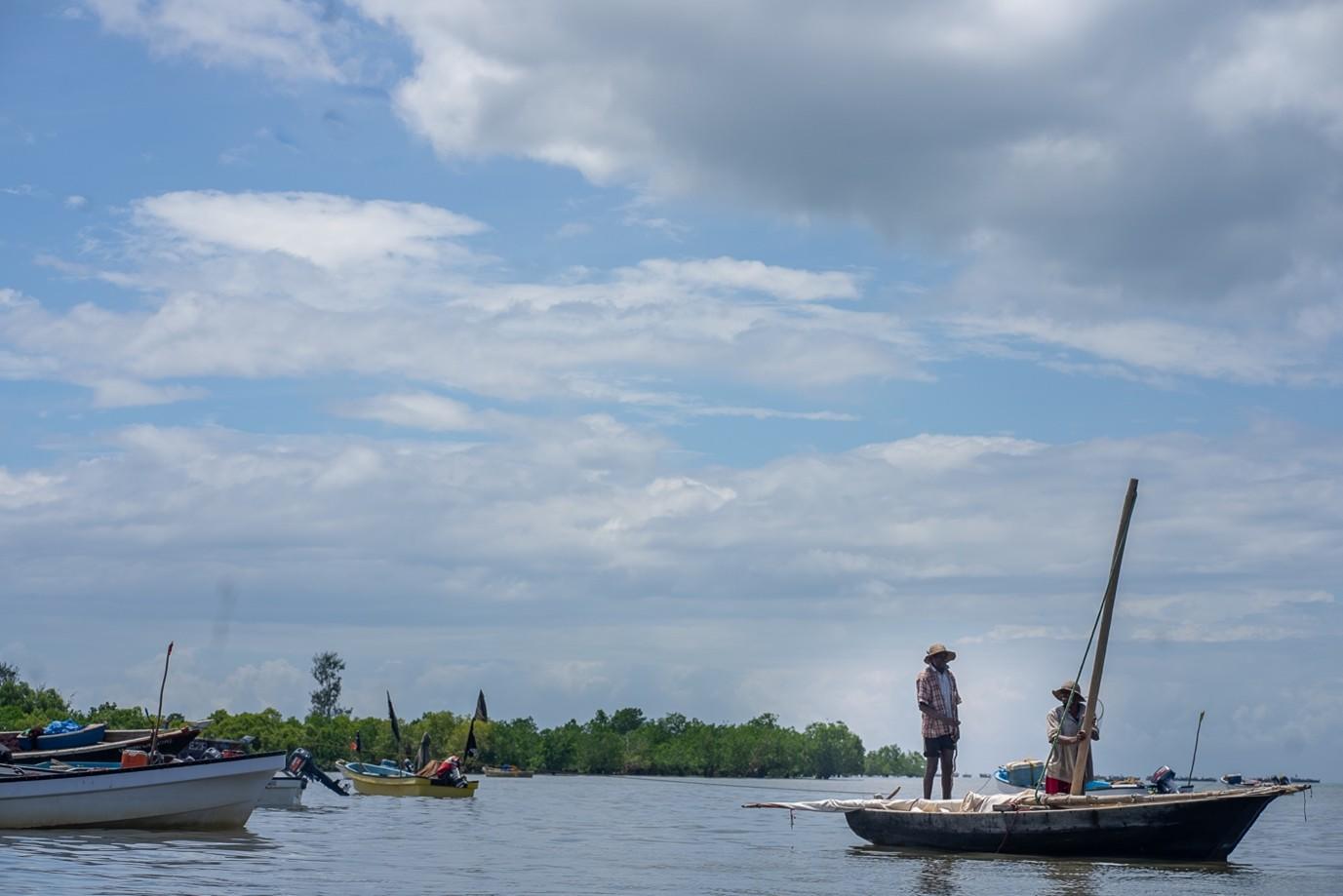 Photo of boats on a calm lake; a person stands on the bow of a sailboat under cloudy skies.