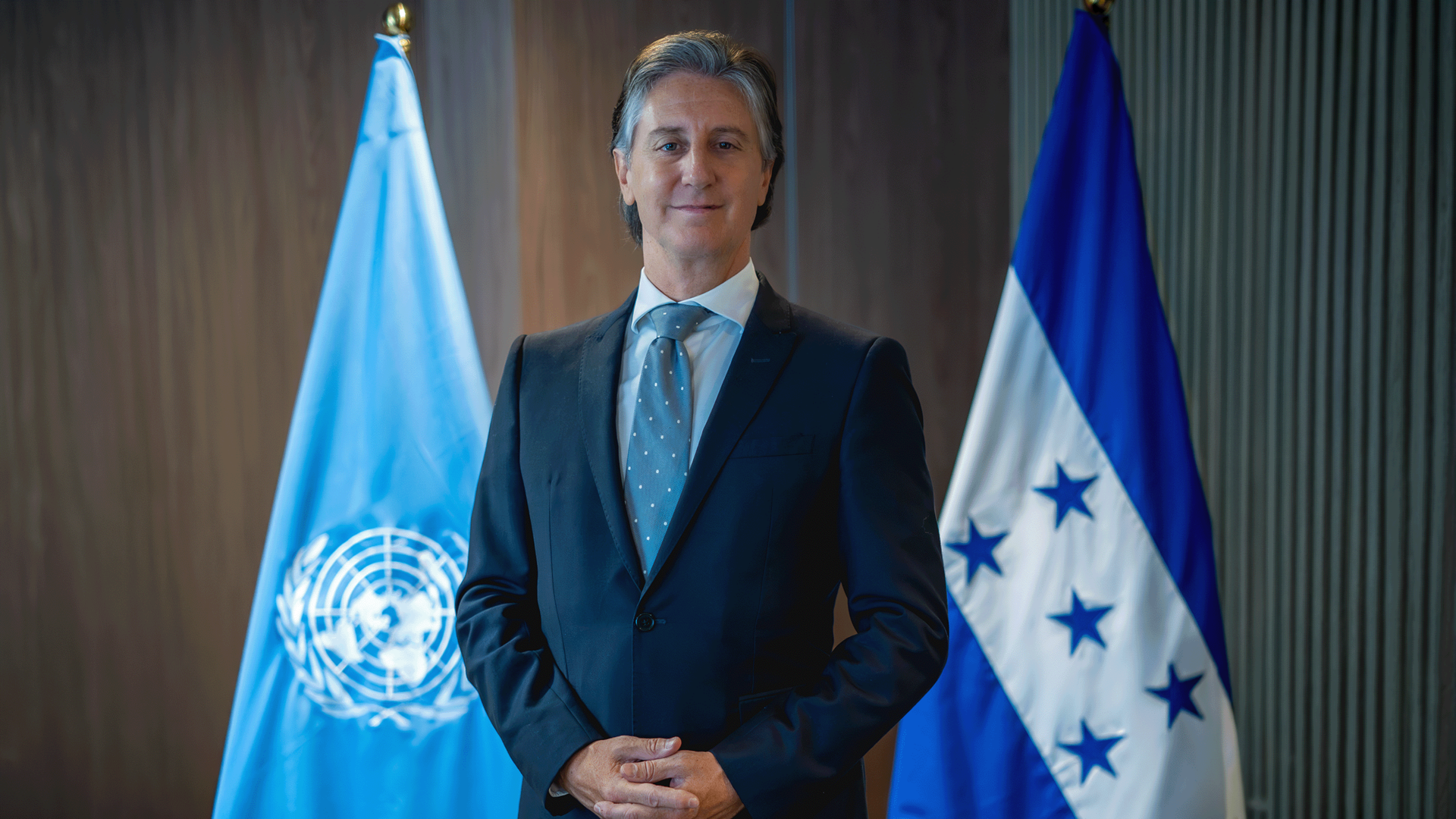 Man in dark suit with blue tie stands with hands clasped, beside UN flag and blue-white flag.