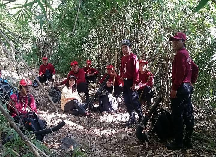 Group of people in red shirts gathered in a wooded clearing, some seated on the ground.