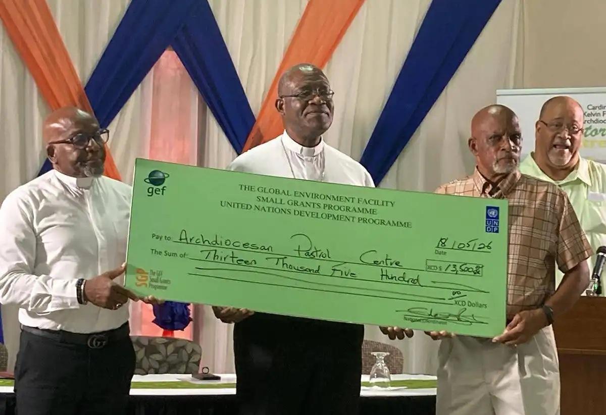 Three men on a stage hold a large green ceremonial check with fourth man in background.