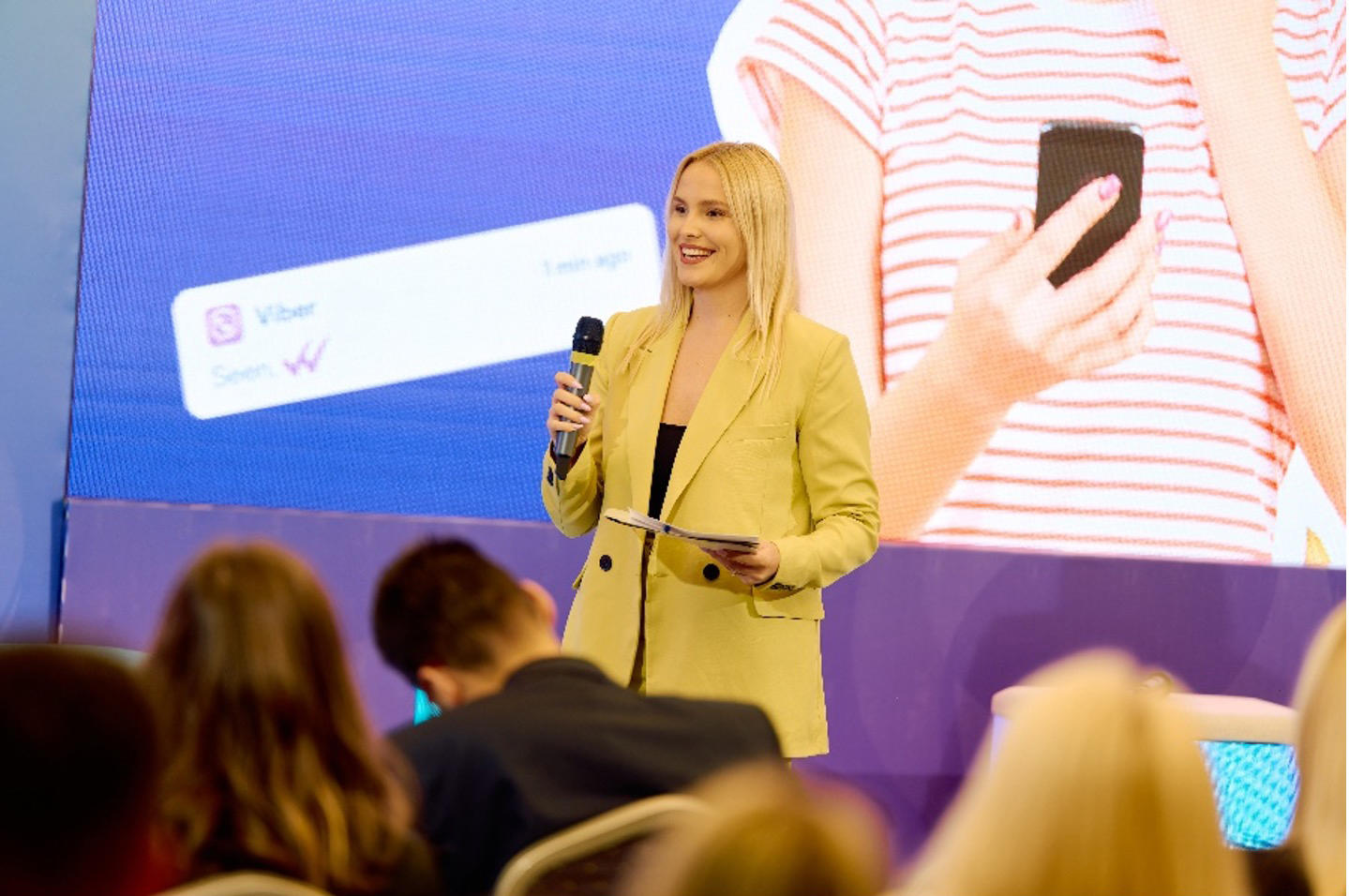 Blonde woman in a beige blazer speaks into a microphone on stage, audience in foreground.