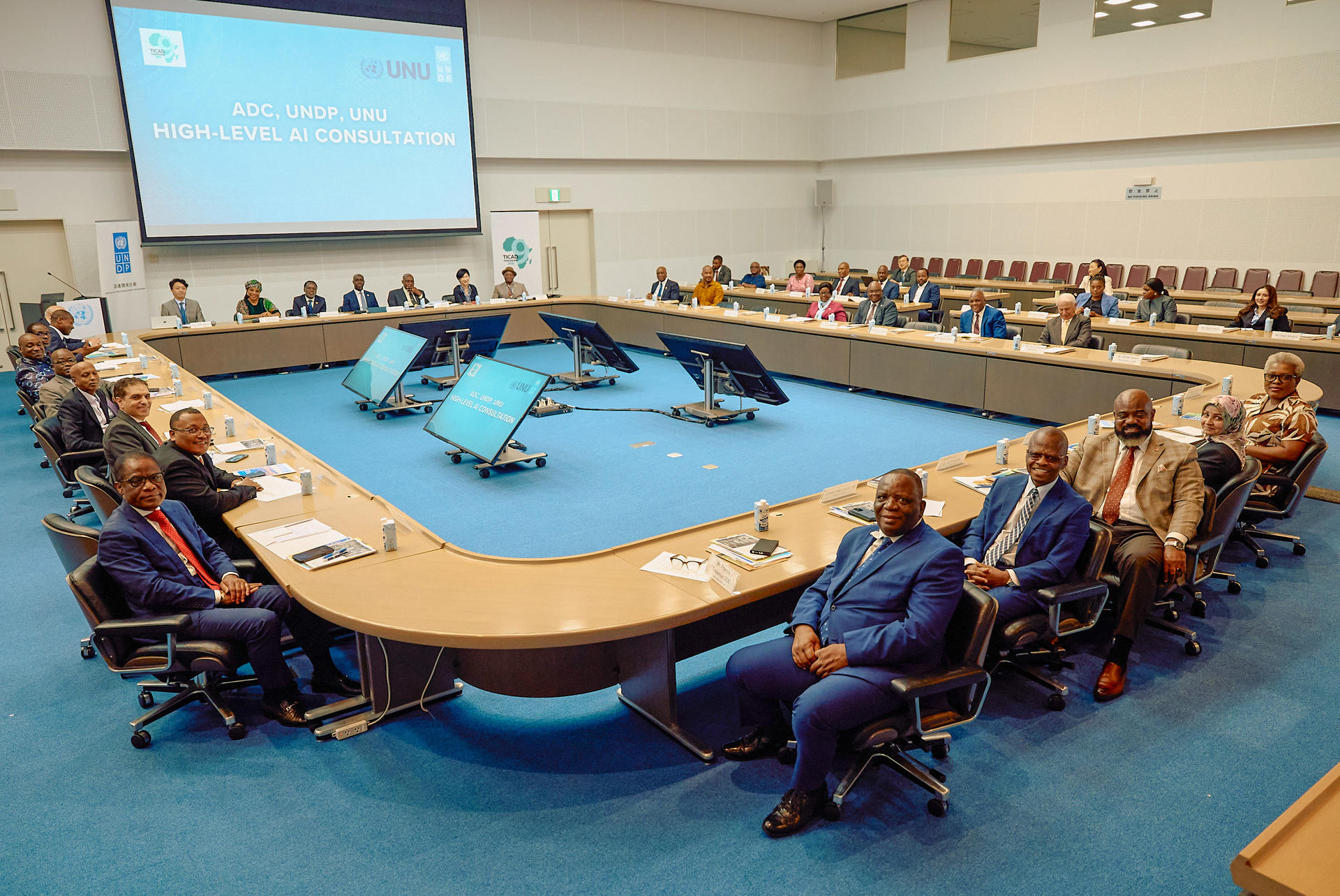 Boardroom meeting with attendees around a U-shaped table; presentation on screen.