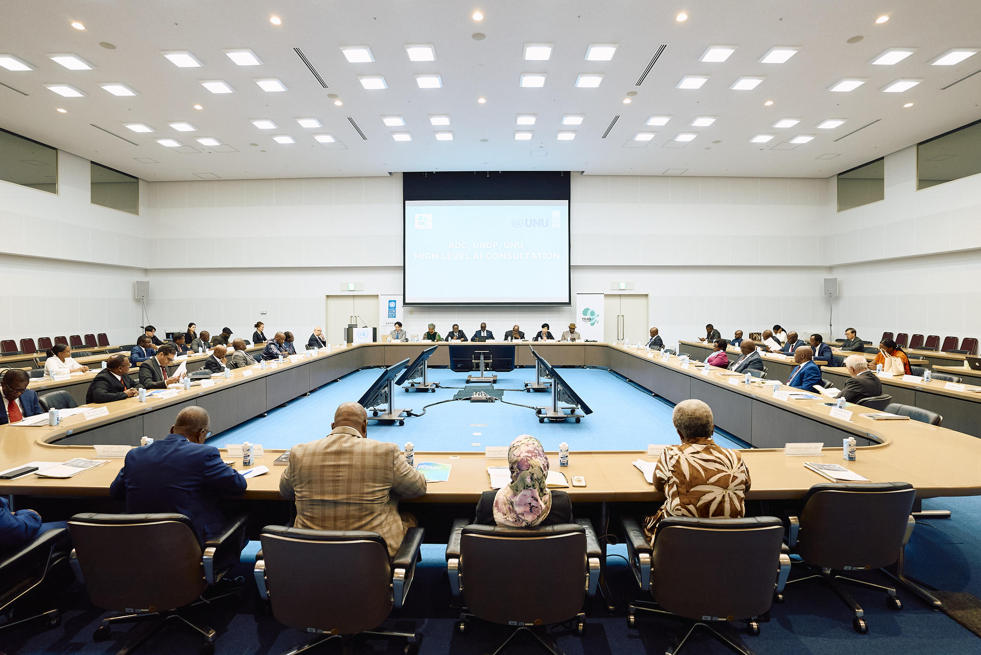 Large conference room with attendees around rectangular table and a projection screen at the front.