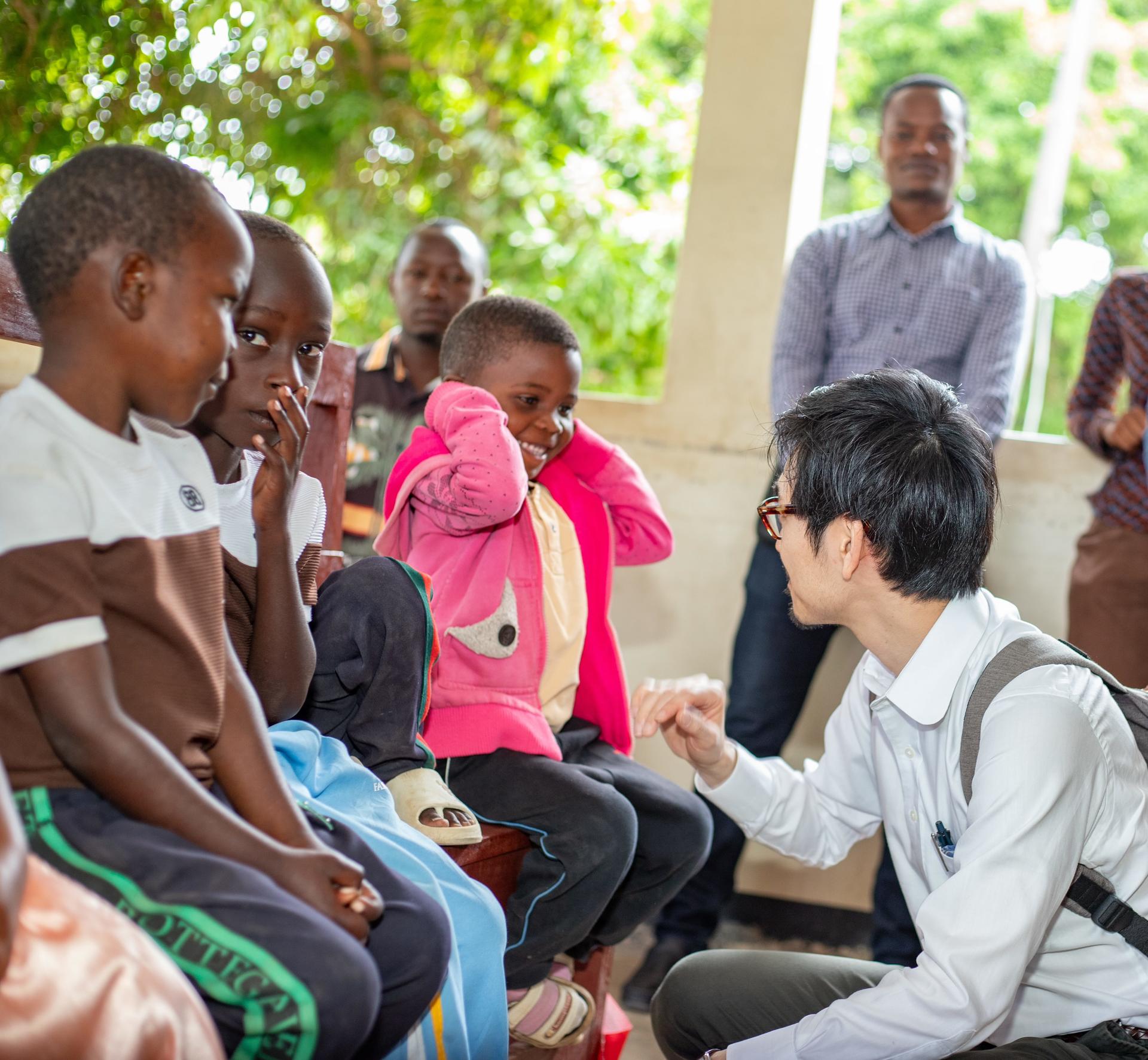 Male volunteer kneels and talks with a group of children outdoors under shady green trees.