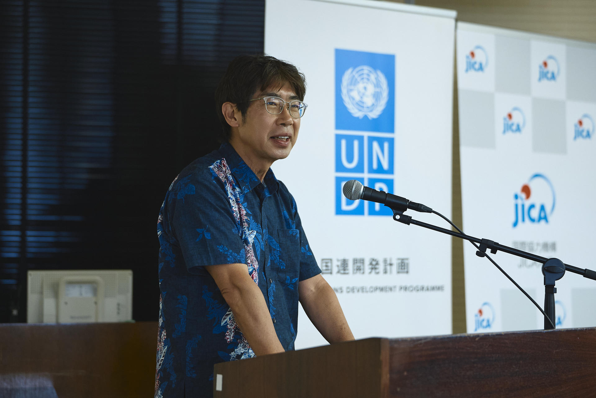 Speaker at podium wearing a blue shirt; backdrop features UN and JICA logos.