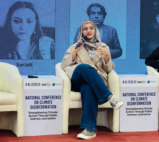 Woman panelist seated on a conference stage with banners and name placards.
