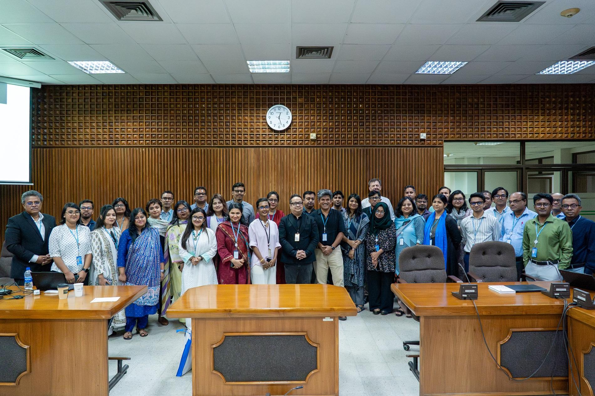 Group photo of adults standing behind desks in a wood-paneled conference room with a clock.