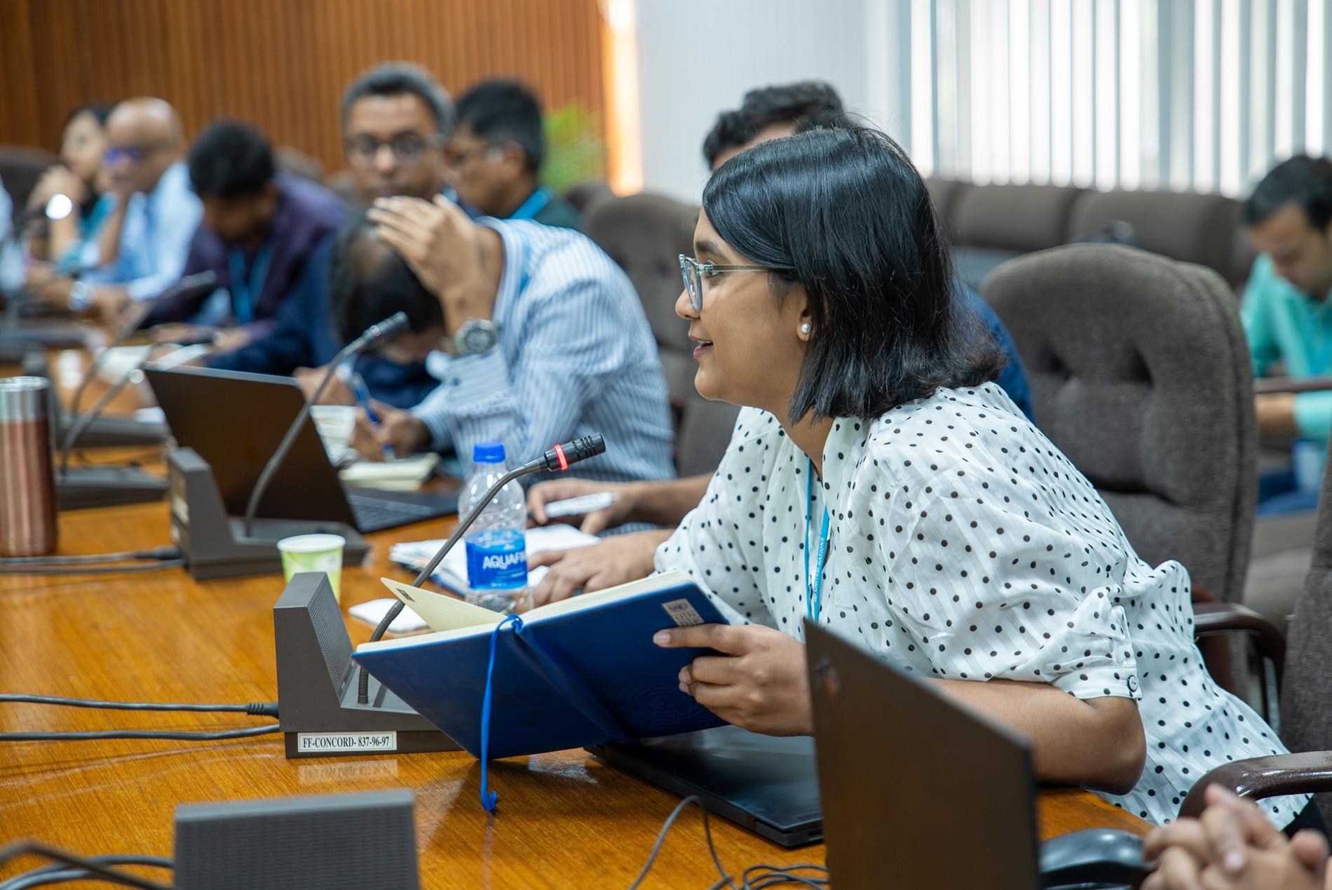 Conference room with attendees; foreground woman in a patterned blouse reads a blue folder.