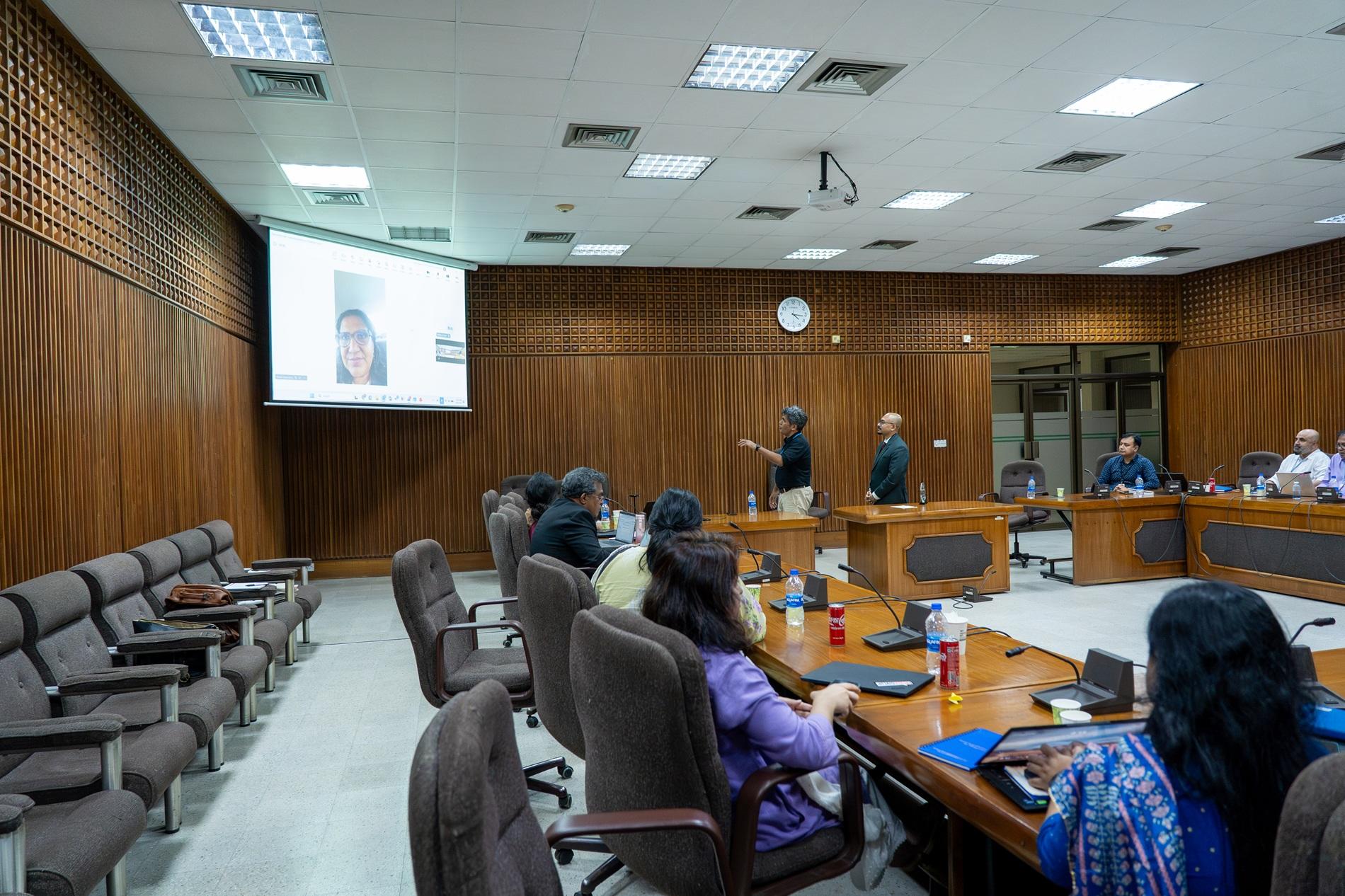 Formal meeting in a wood-paneled conference room, speaker at podium, presentation on screen.