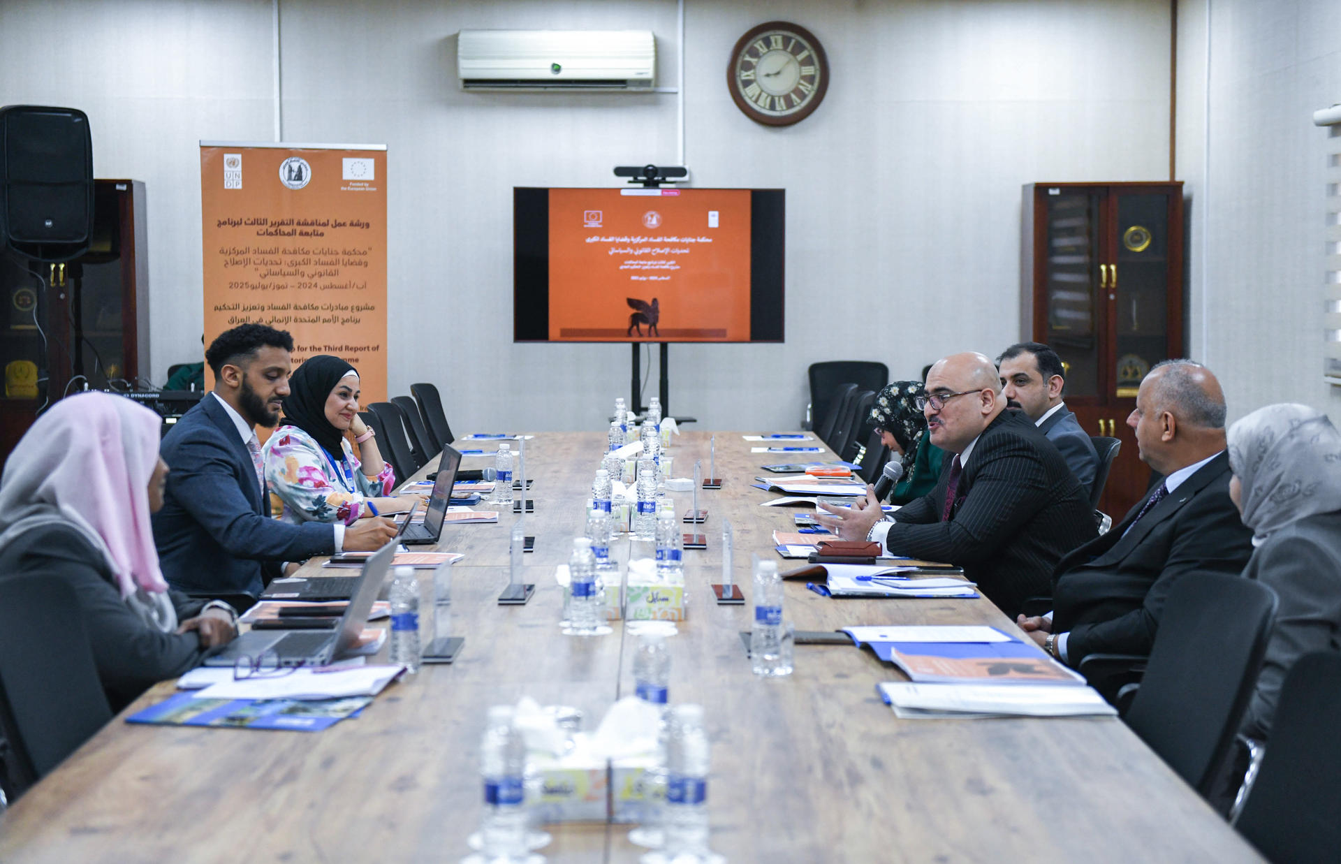 Attendees around a long conference table in a meeting room with a screen at the far end.