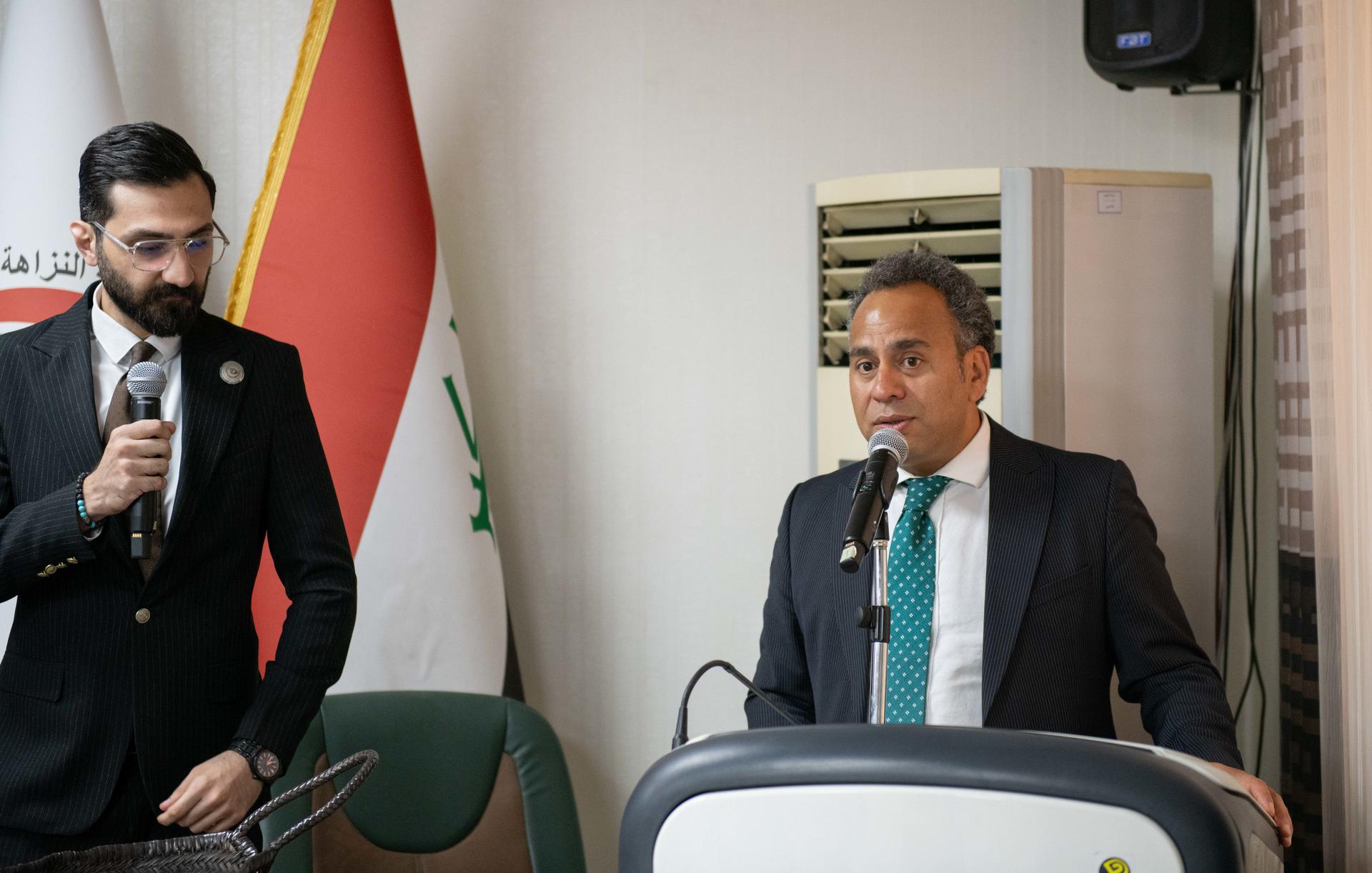Two men in suits at a conference table; one speaks into a microphone; a flag stands nearby.