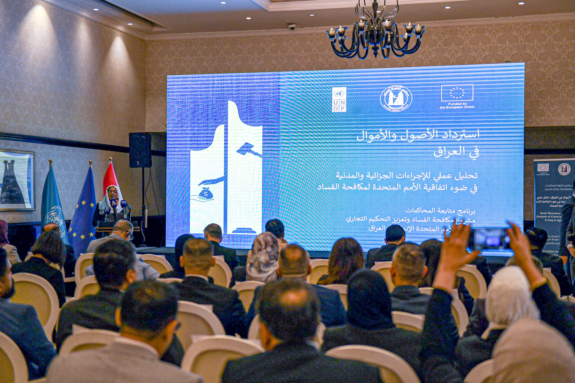 Audience seated in a conference hall watching a large blue presentation screen with logos.