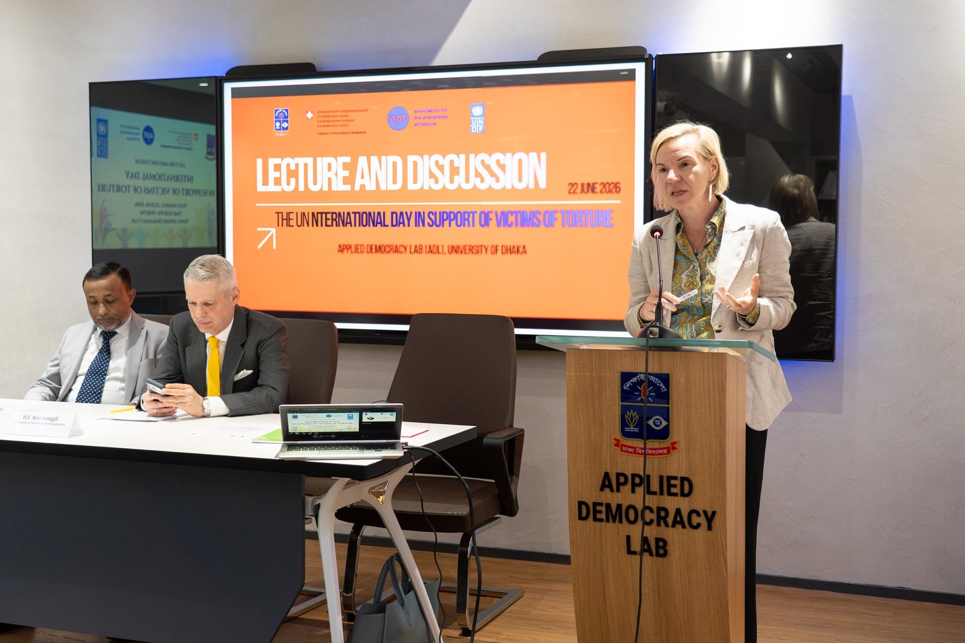 Speaker at podium during panel discussion; two panelists at table, orange slide on screen.