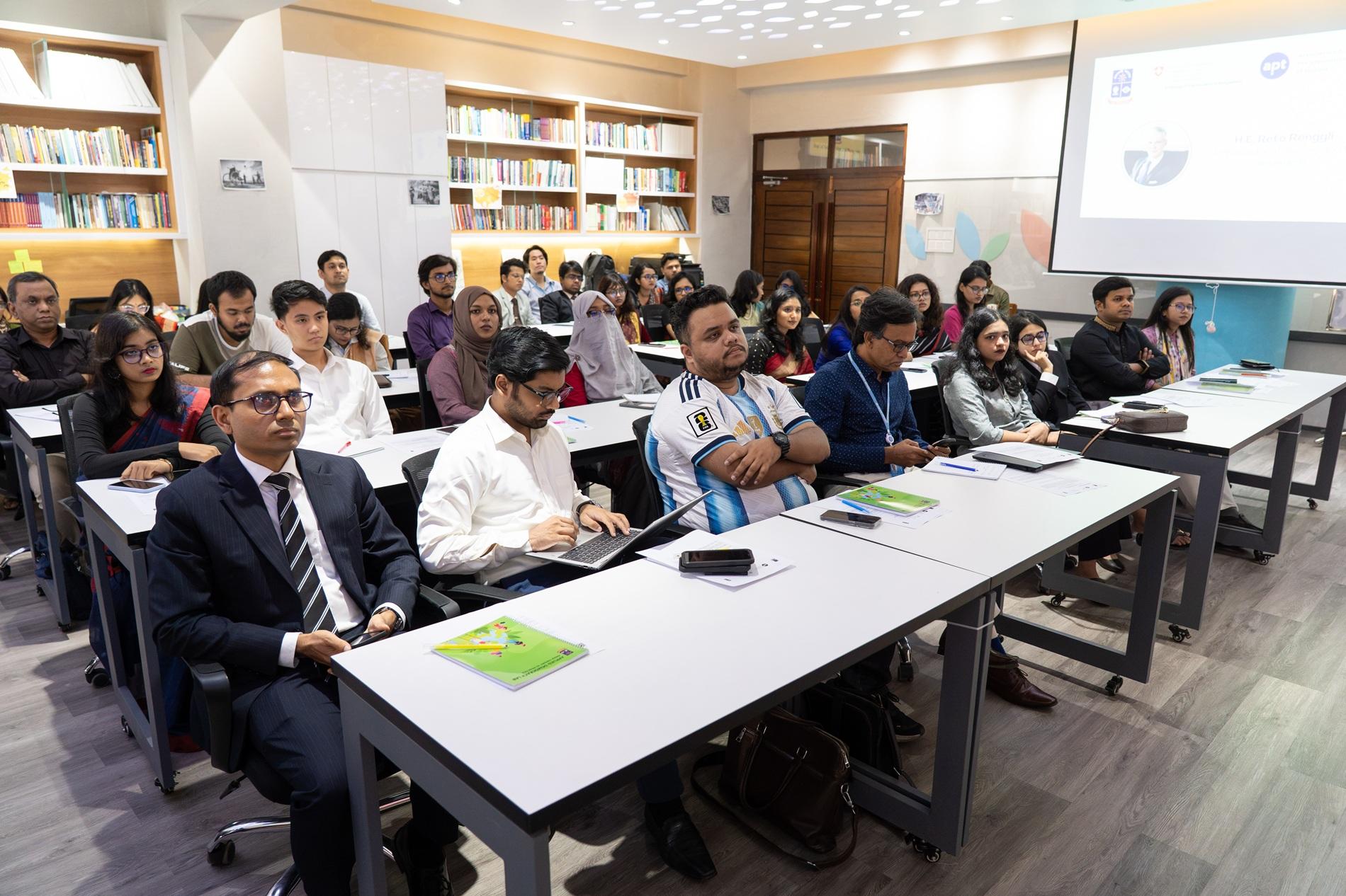 Group of people seated at desks in a classroom; projector screen at front and bookshelves on walls.