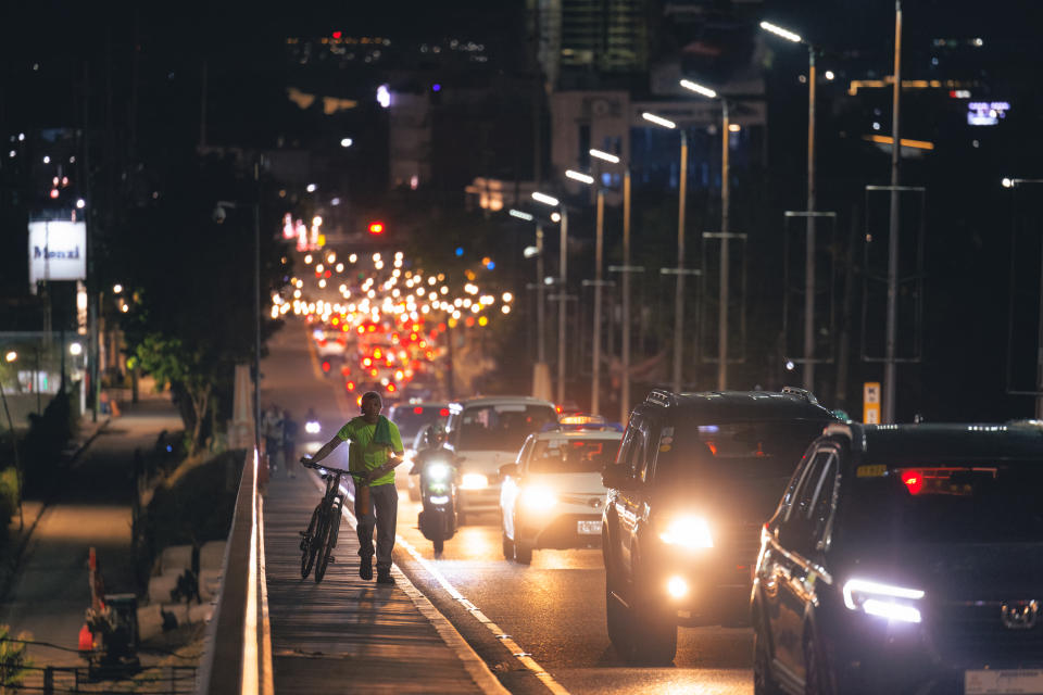 Night city street with cars in traffic and two people on a sidewalk as fireworks light the sky.