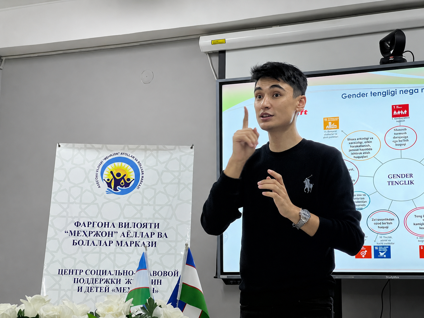 Man speaks into a microphone during a presentation beside a banner and projection screen.
