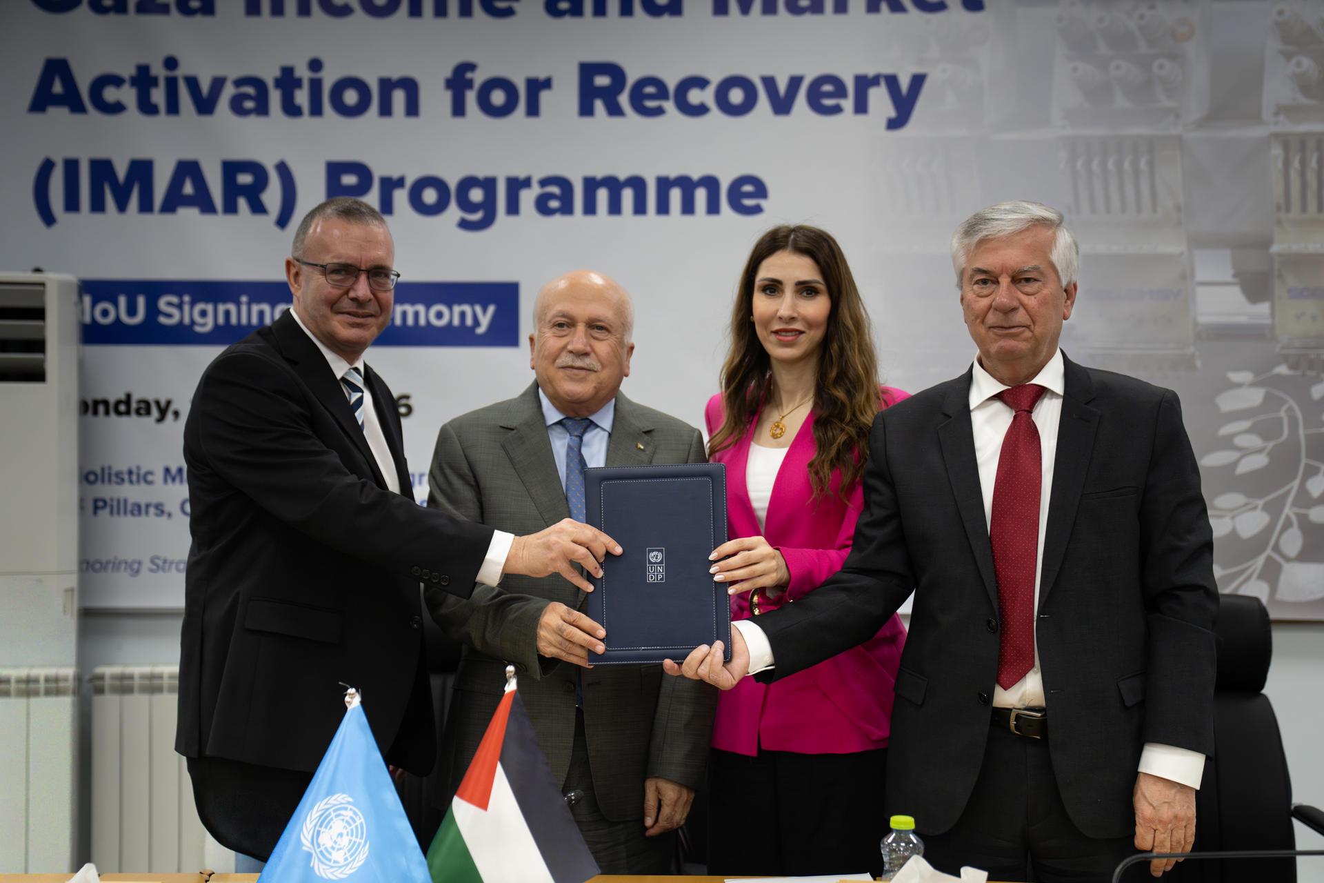 A man hands a folder to a woman in a pink blazer at a formal event, with other officials nearby.