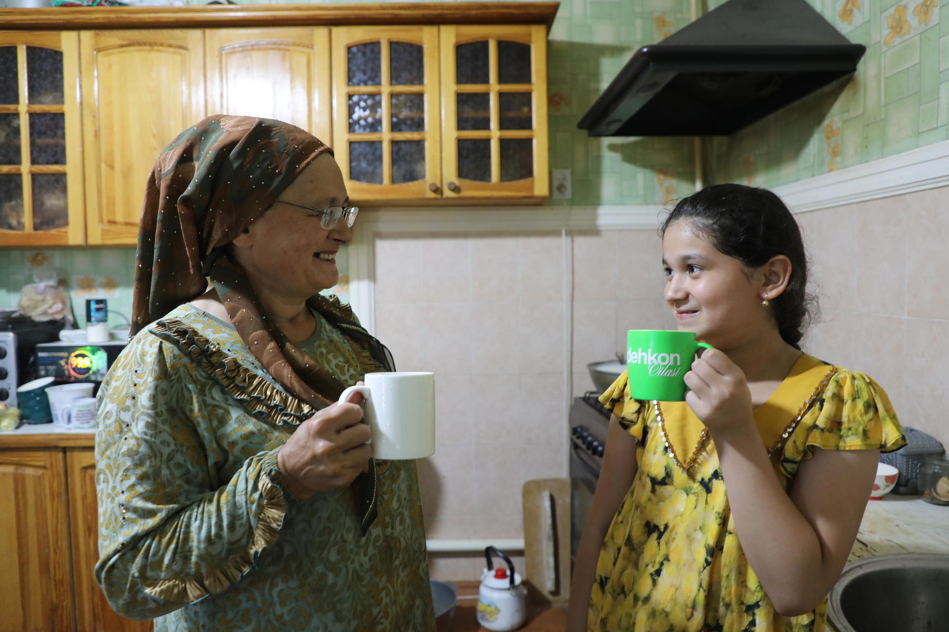 Two women in a kitchen, one in a patterned dress with headscarf, the other in yellow, sipping mugs.