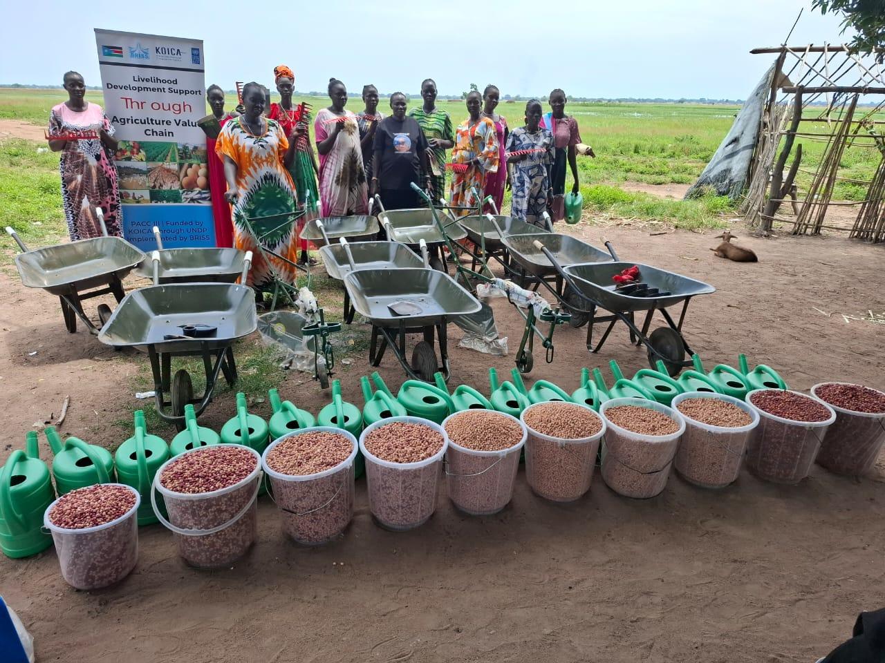 Group of people at a garden with rows of potted seedlings and a sign in the background.