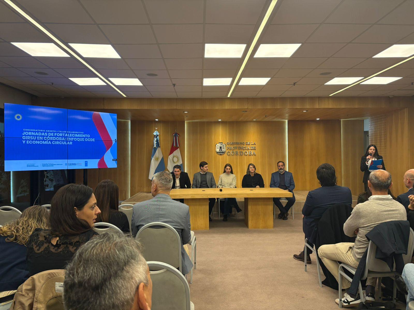 Panelists seated at a long table on a stage in a wood-paneled conference room; audience listening.