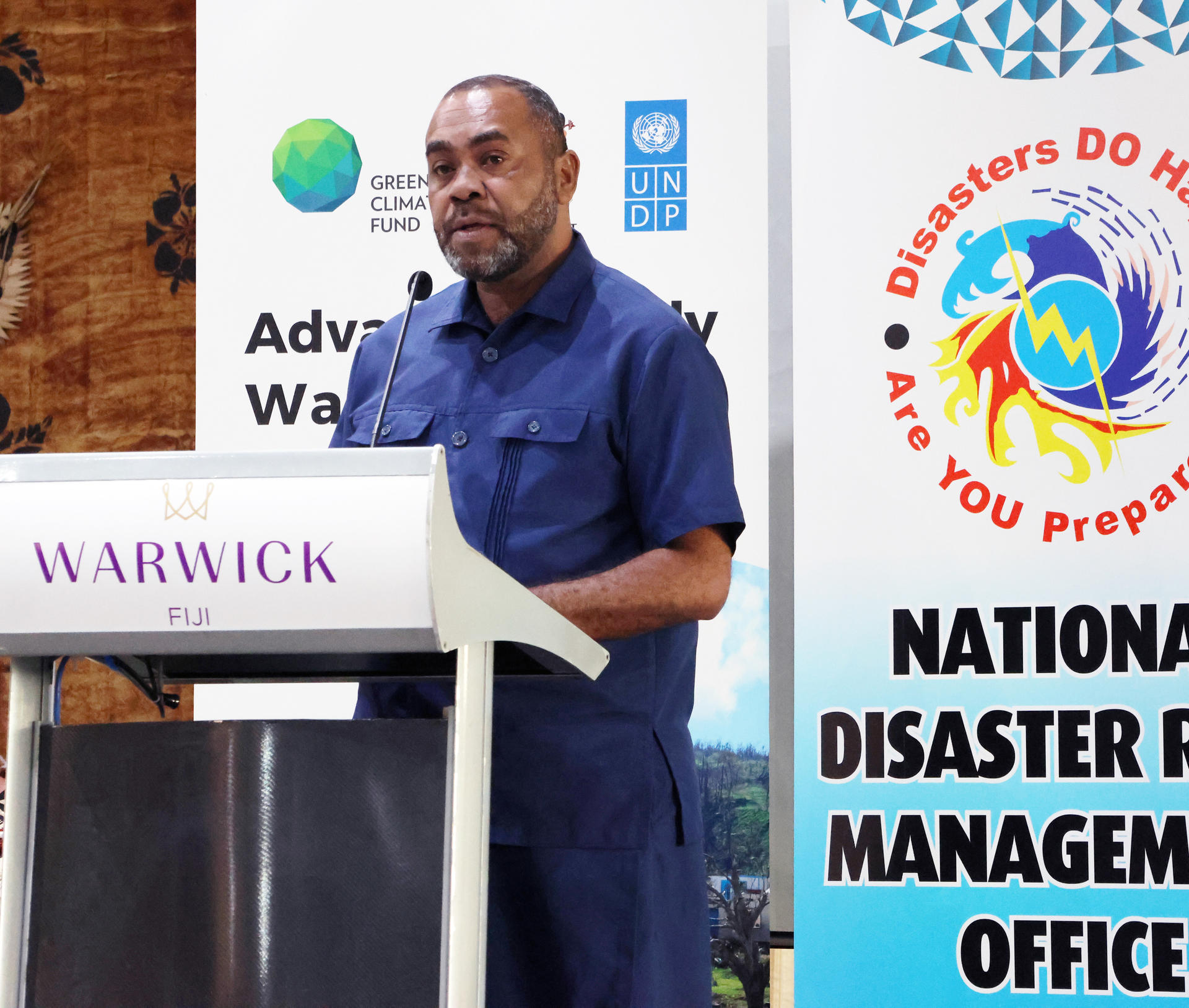 Man in blue shirt speaks at Warwick podium during disaster management conference.