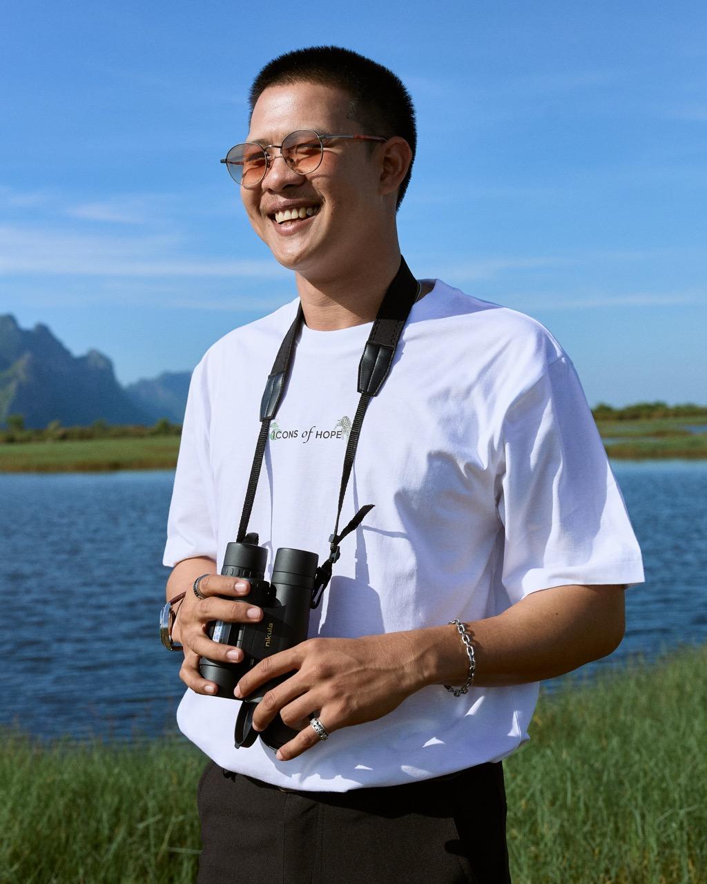 Smiling man outdoors by a lake with mountains, holding binoculars.