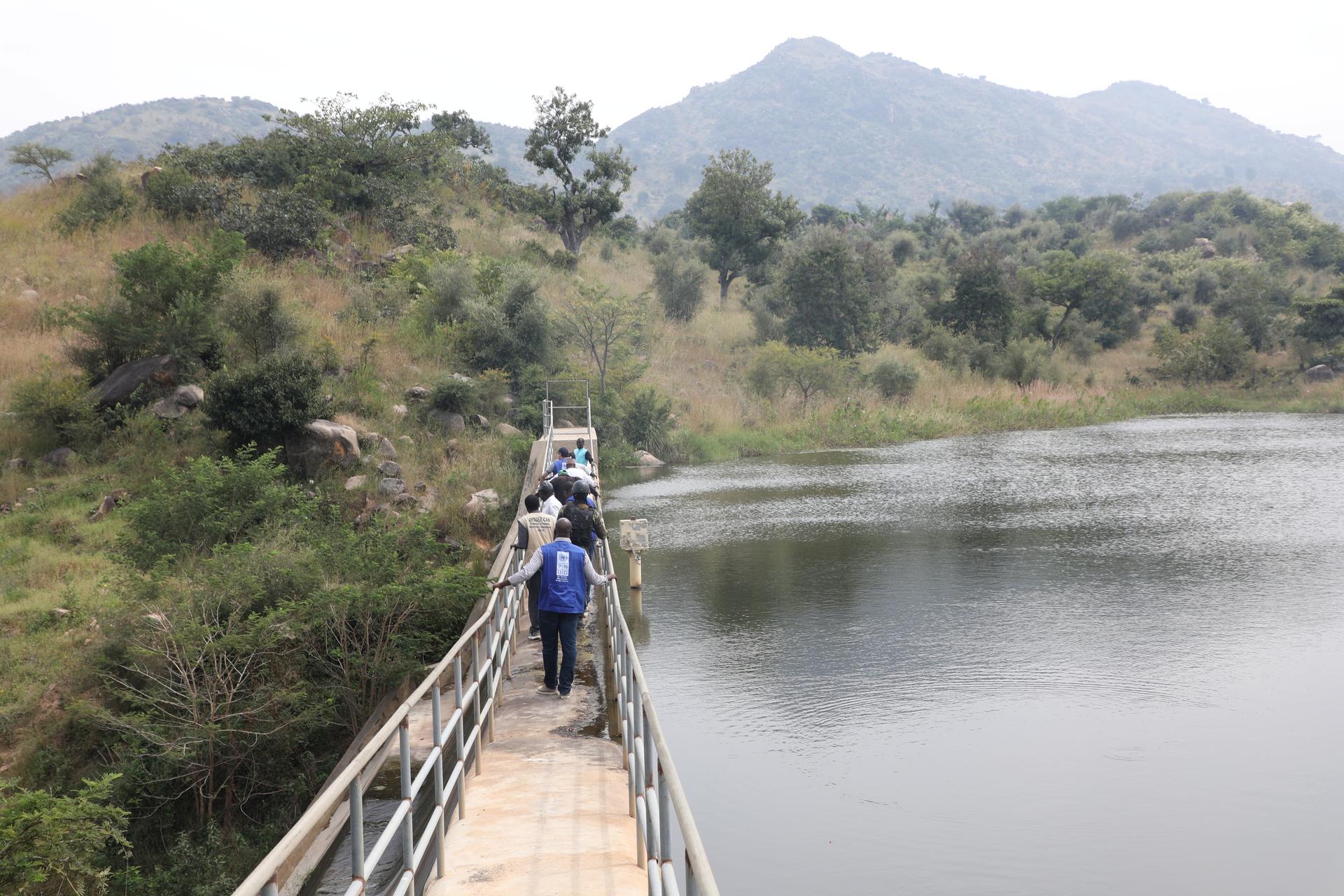 Pedestrian bridge over a calm lake with hikers walking; hills and trees in the distance.