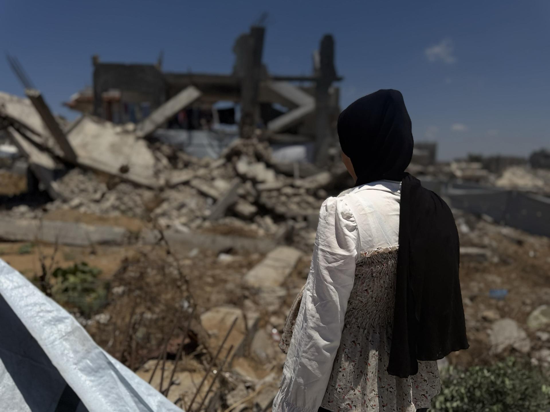 Person in white shirt and black headscarf stands amid rubble of a destroyed building.