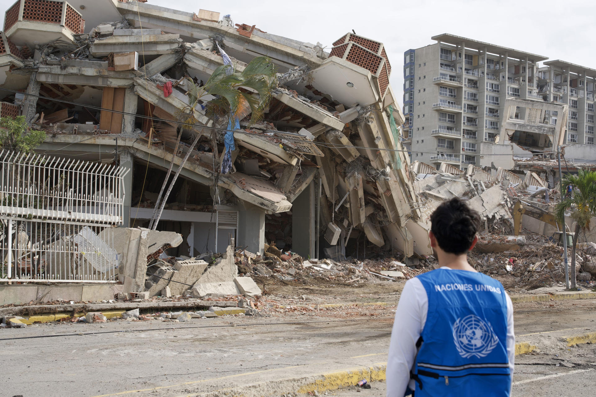 Person in a blue safety vest surveys a collapsed multi-story building and rubble.