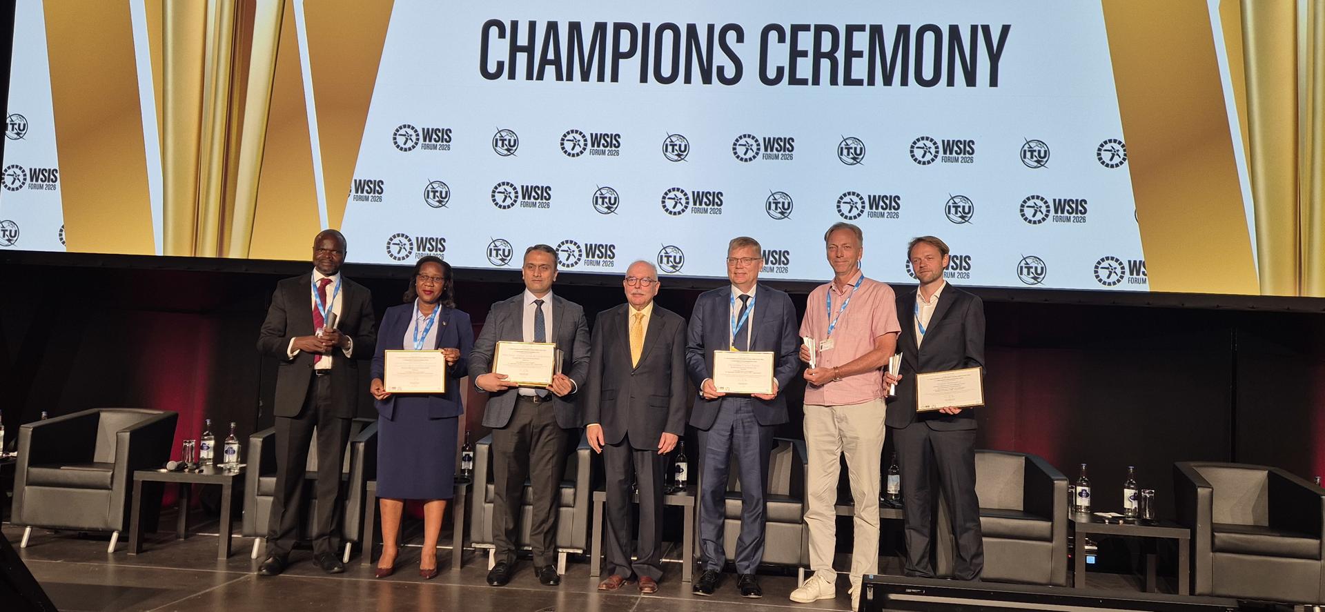 Group of people on stage receiving certificates at the Champions Ceremony, gold backdrop.