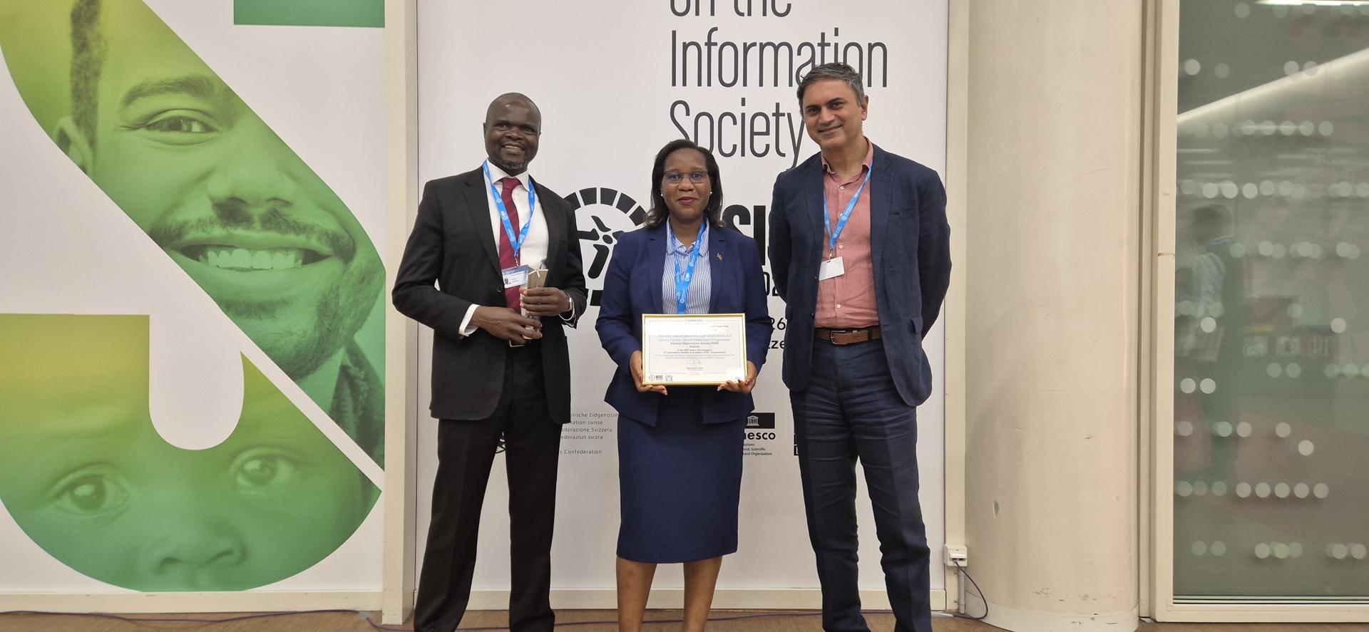 Three people in formal attire pose in a hallway; center holds a certificate, banner behind.