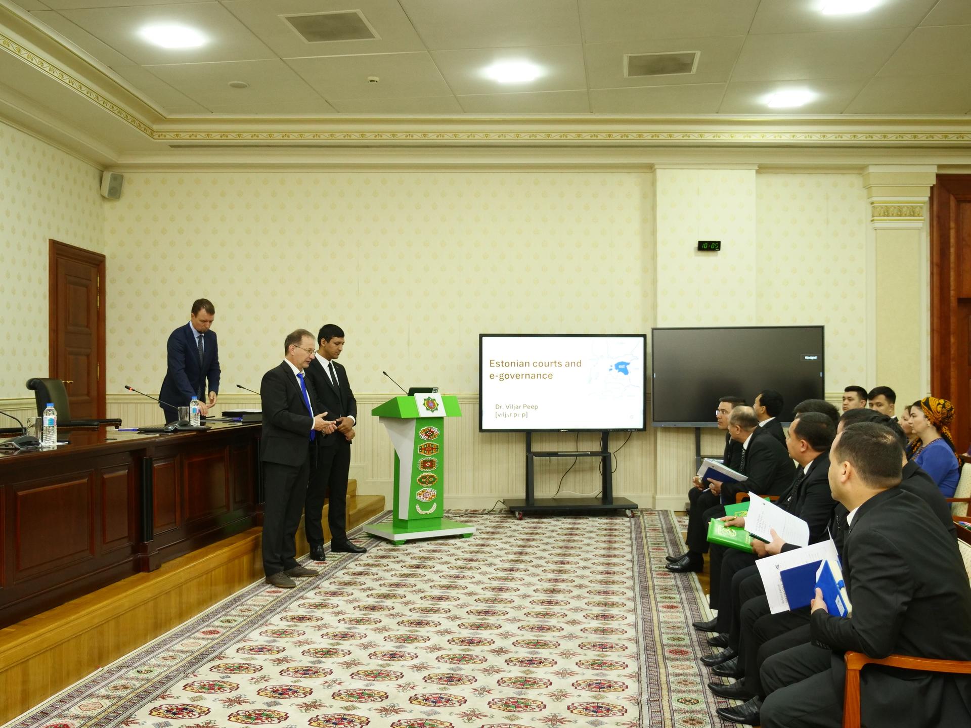Photograph of a formal conference room with a panel on the left and audience on the right.