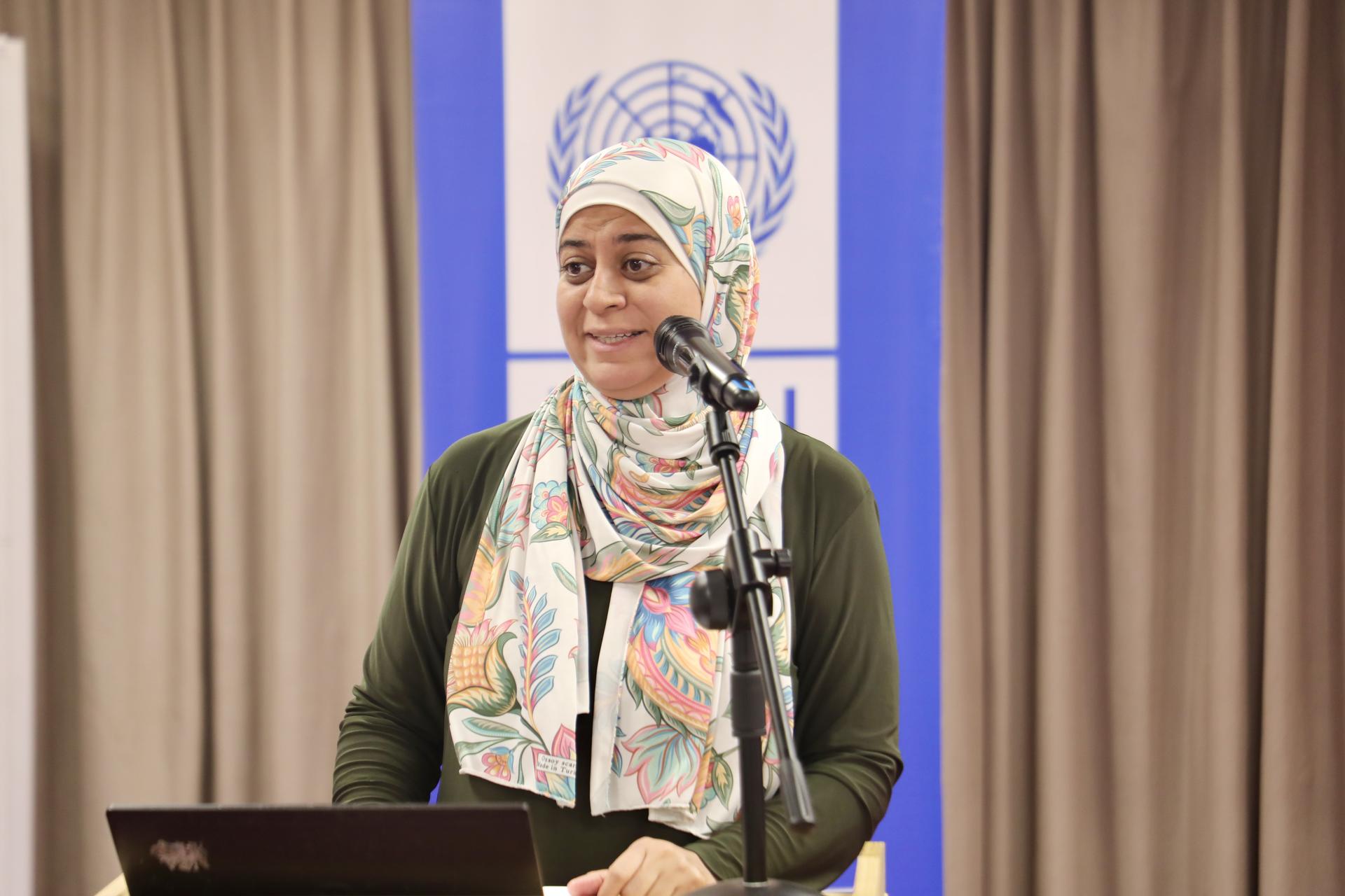 Photograph: Woman in a patterned hijab at a podium with a microphone, UN banner behind.