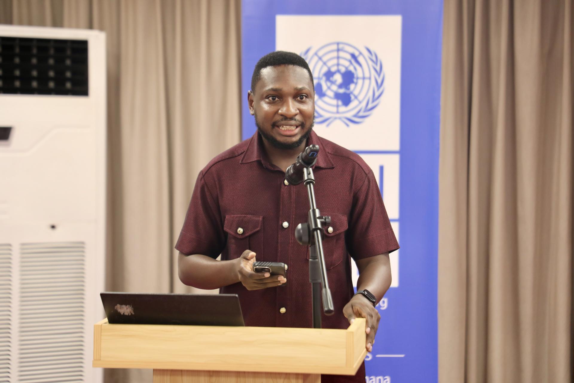 Man in a maroon shirt speaks at a podium with a UN banner behind him.