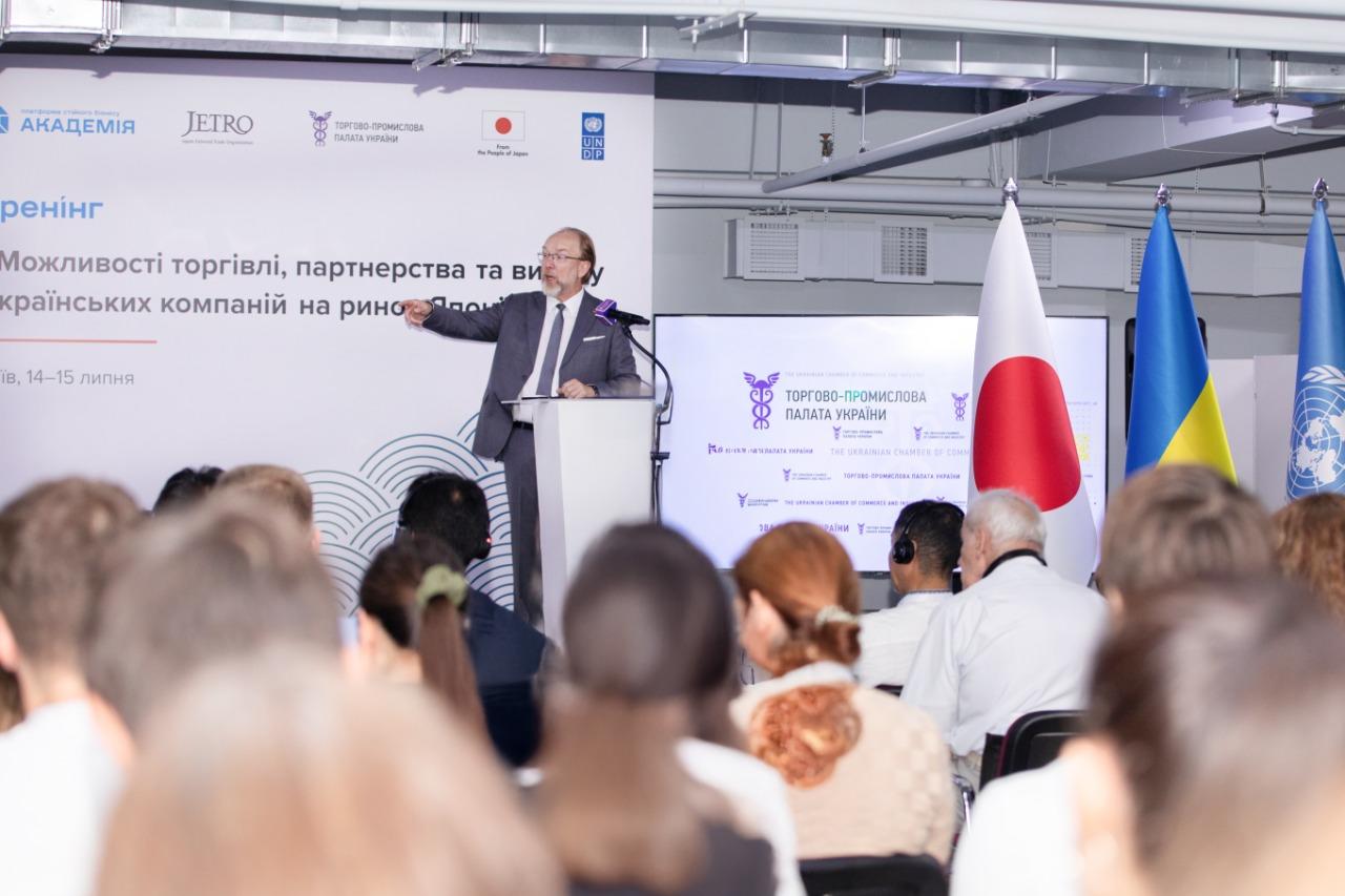 Conference speaker at podium addressing audience; banners and Japanese flag visible.