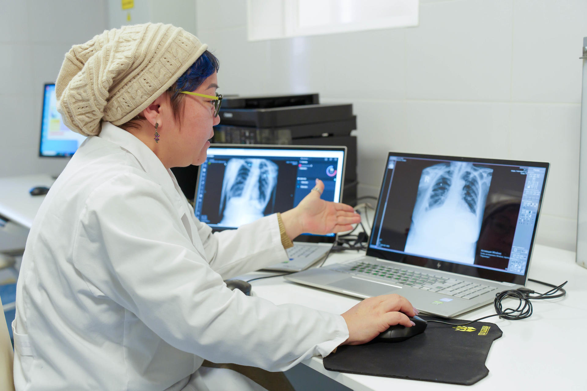 Photo of a radiology technician in a white coat and hairnet reviewing X-ray images on dual monitors.