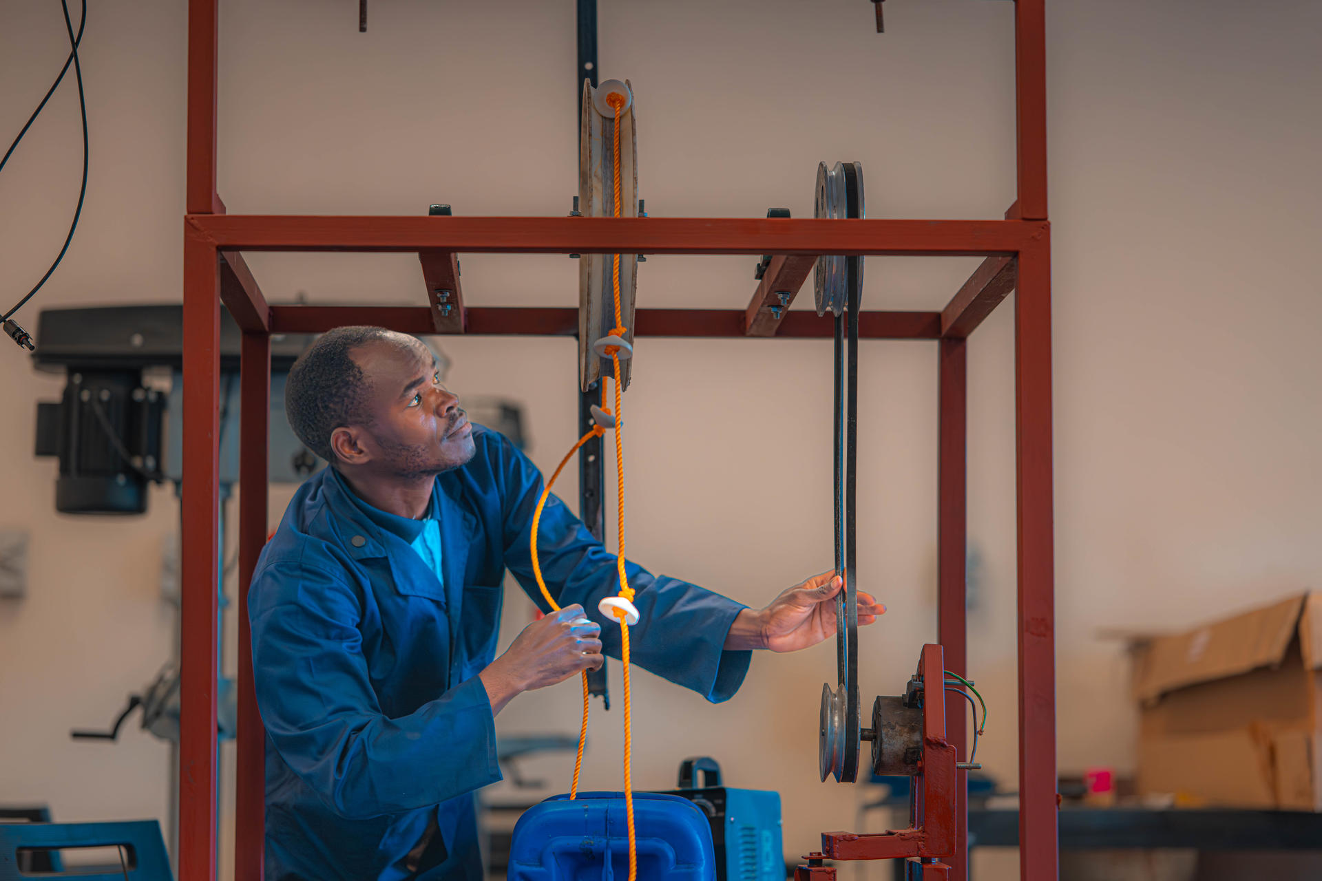Man in a blue jacket uses a tool on a metal red-framed structure inside a workshop.