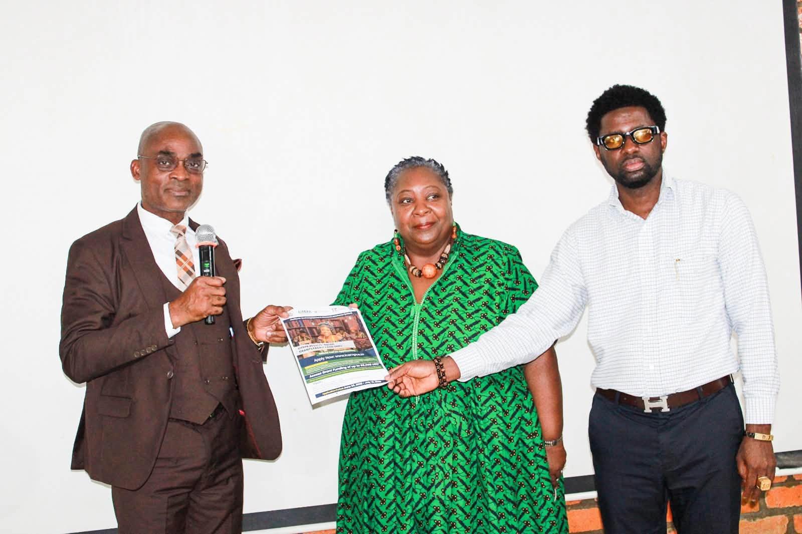 Three people on stage: man in brown suit, woman in green with certificate, man in white shirt.