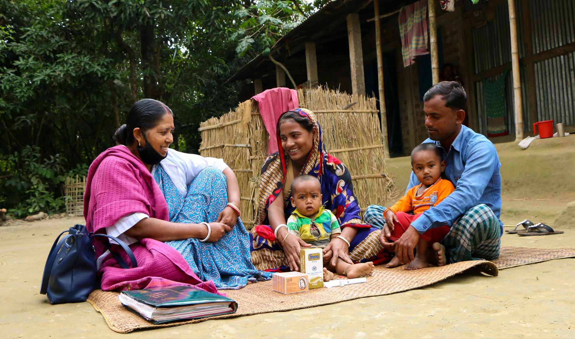Rural family of six sits on a mat outside a bamboo hut; a baby rests on a woman's lap.