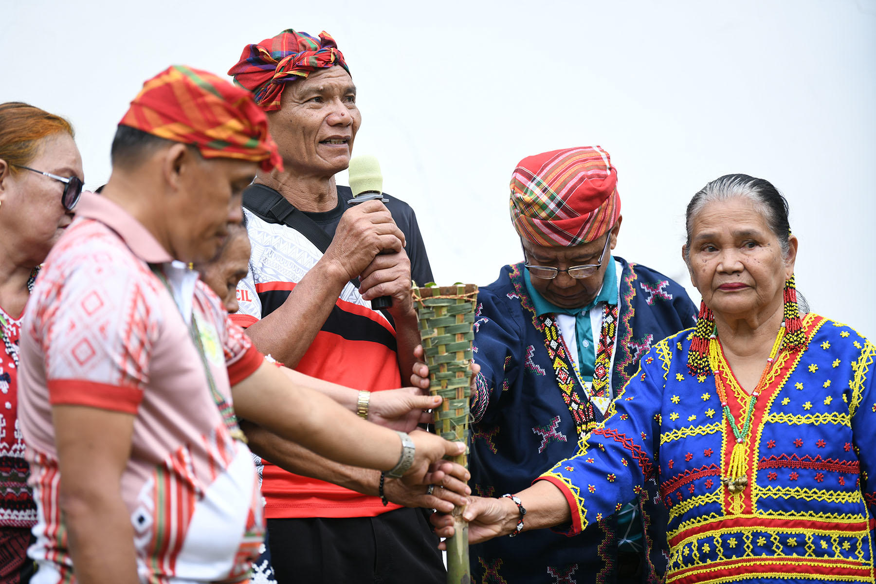 Group of Indigenous people in colorful traditional dress at a ceremony; man speaks into a mic.