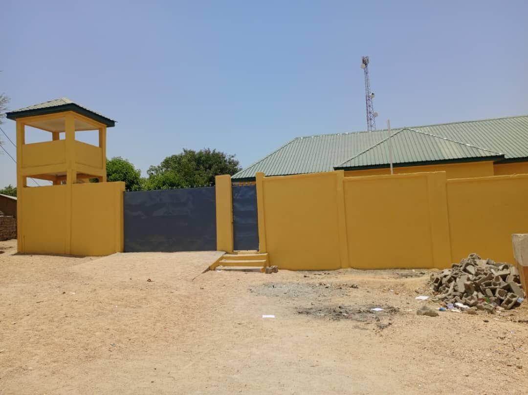 Sunlit yellow-walled compound with blue gate, guard tower, and rubble on sandy ground.