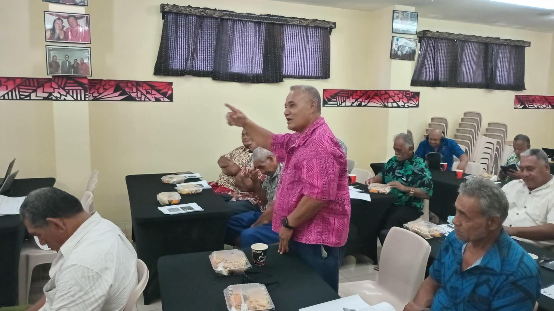 Man in pink shirt stands and gestures to a seated audience in a small conference room.