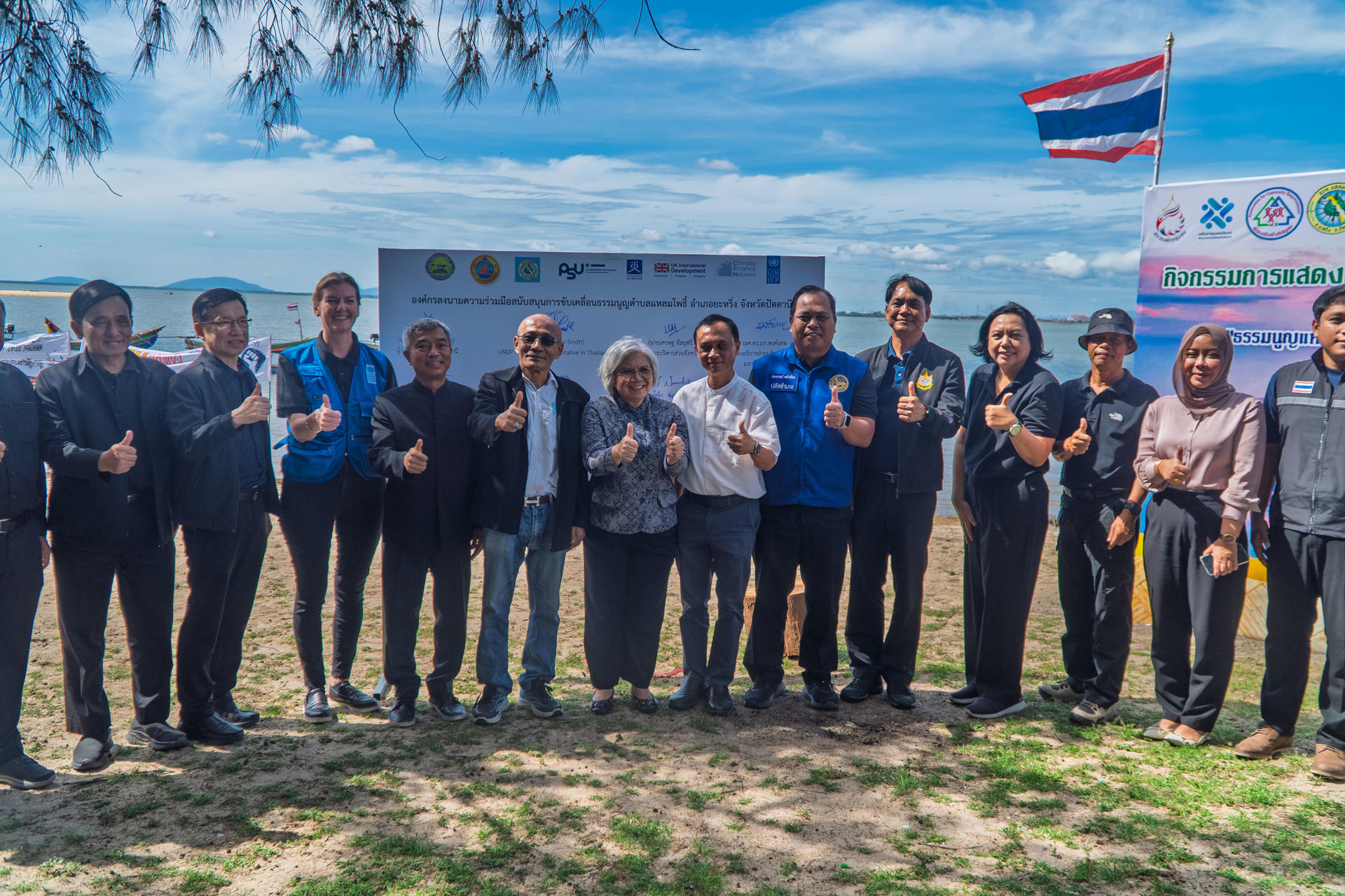 Group of people posing on a sunny beach with a Thai flag and a banner with logos in the background.