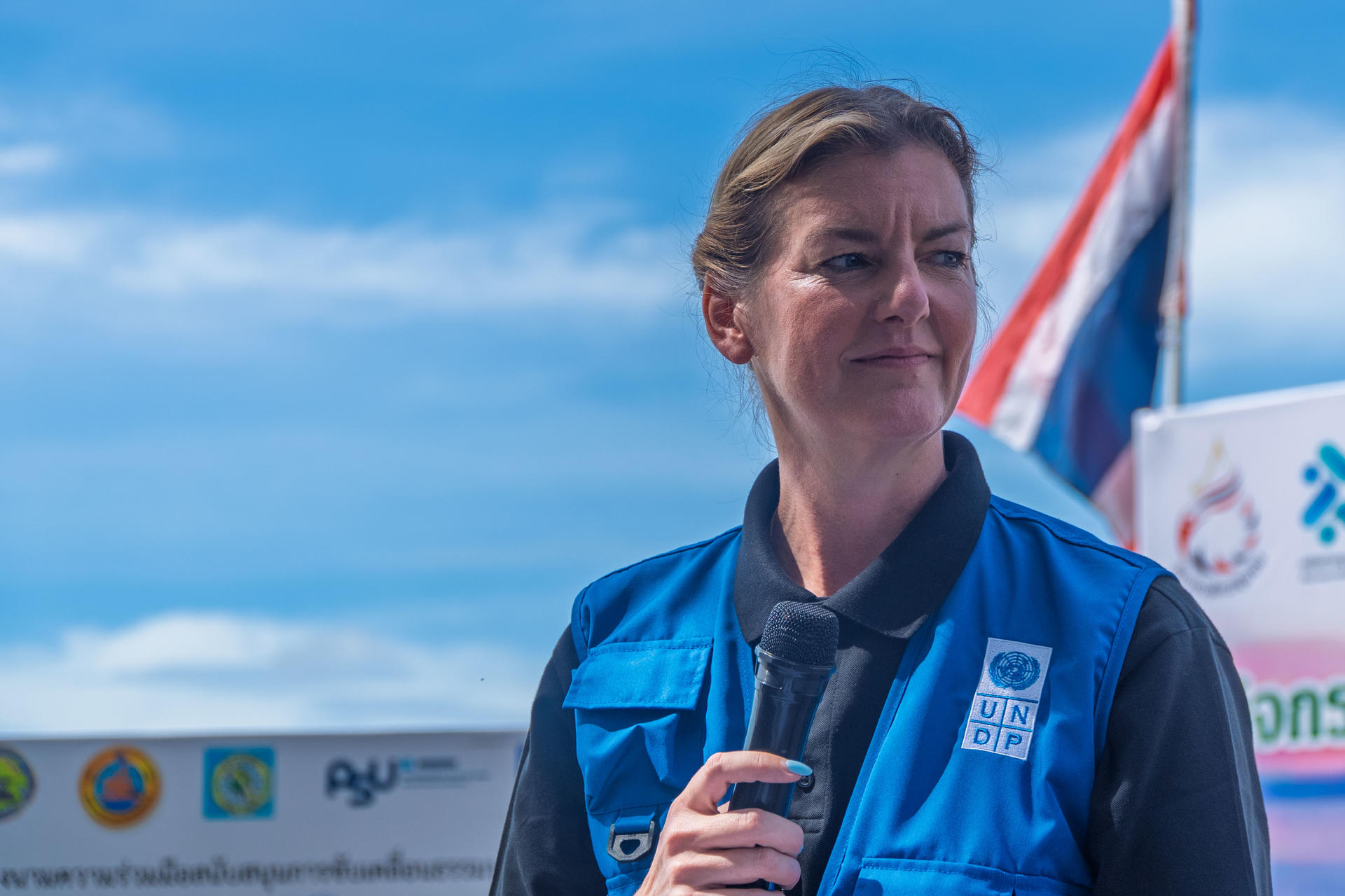 Female presenter in a blue vest speaks into a microphone at an outdoor event.