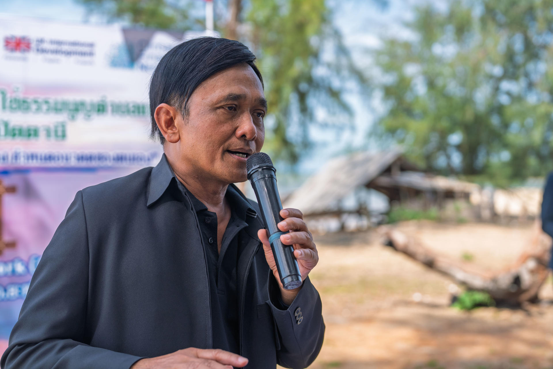 Man in a dark suit speaks into a microphone at an outdoor event, with a banner behind him.