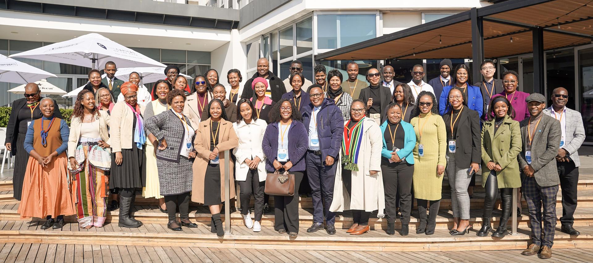 Group photo of diverse adults standing together outdoors in front of a building and tents.