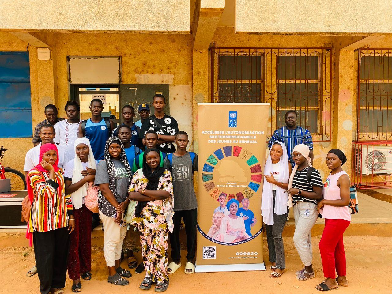 Diverse group of people posing outside a yellow building, with a banner displaying a sun logo.