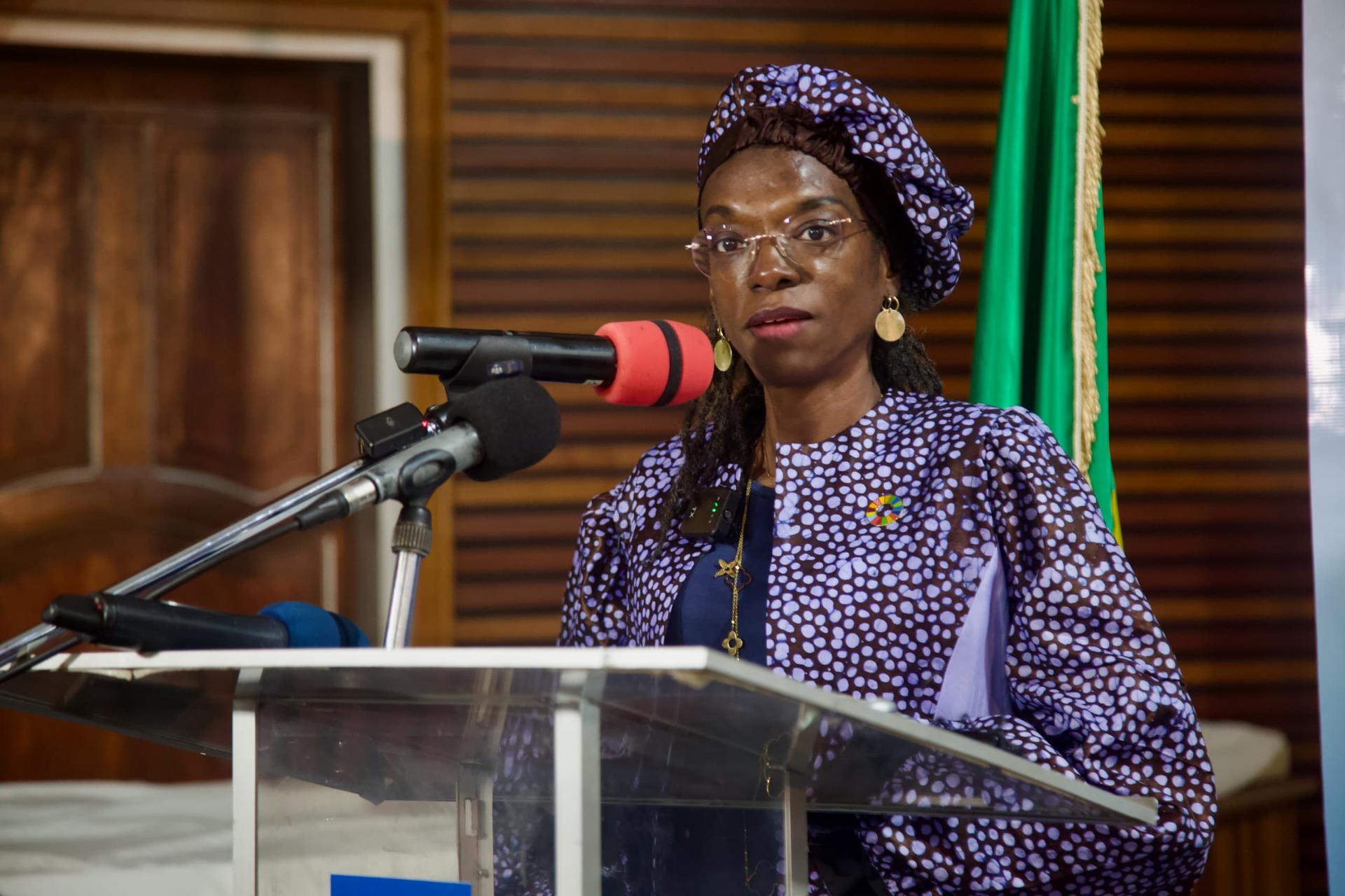 Woman in purple dress and headwrap speaks at a microphone podium, green flag behind.