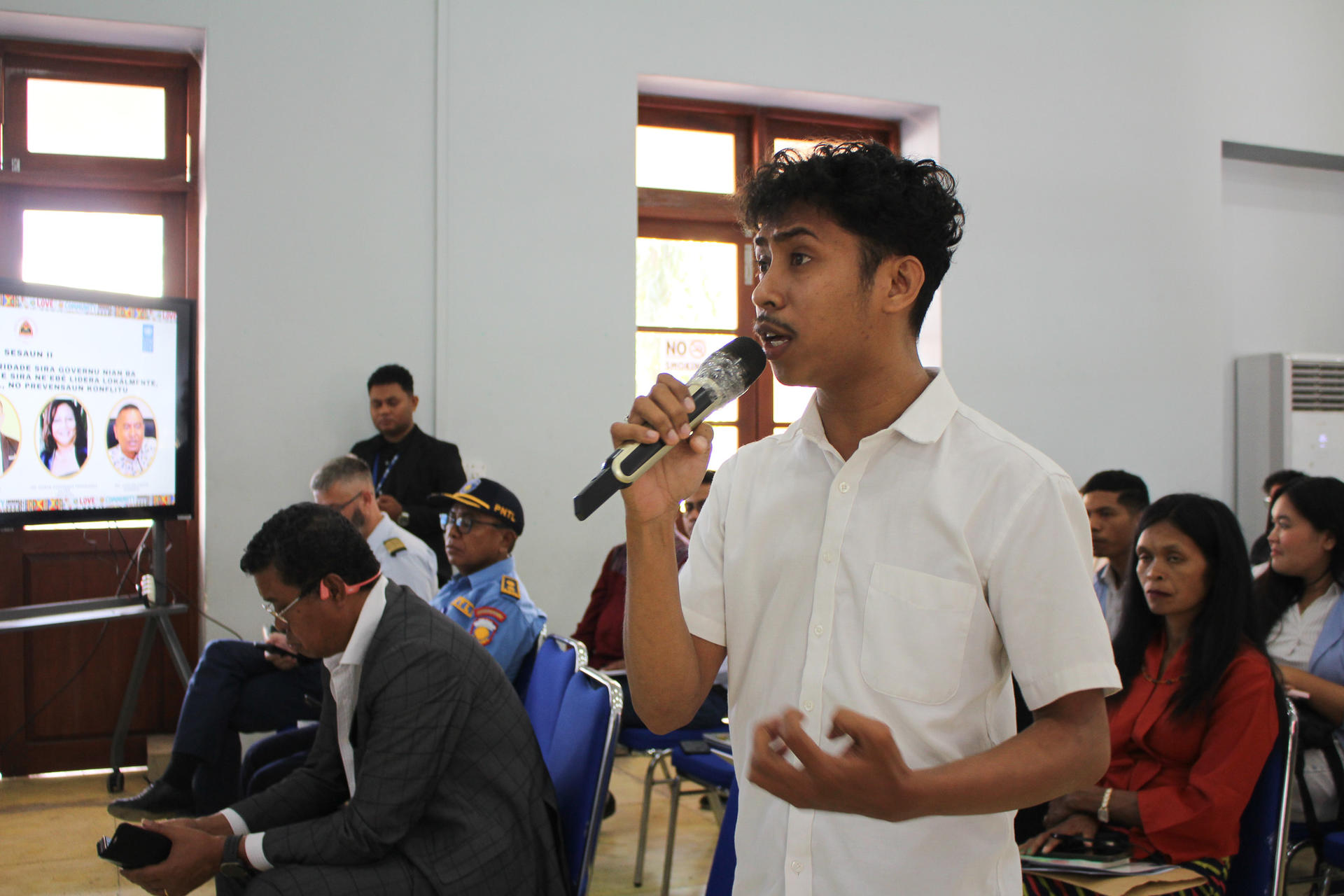 Young man in a white shirt speaks into a microphone during a seminar, with an attentive audience.
