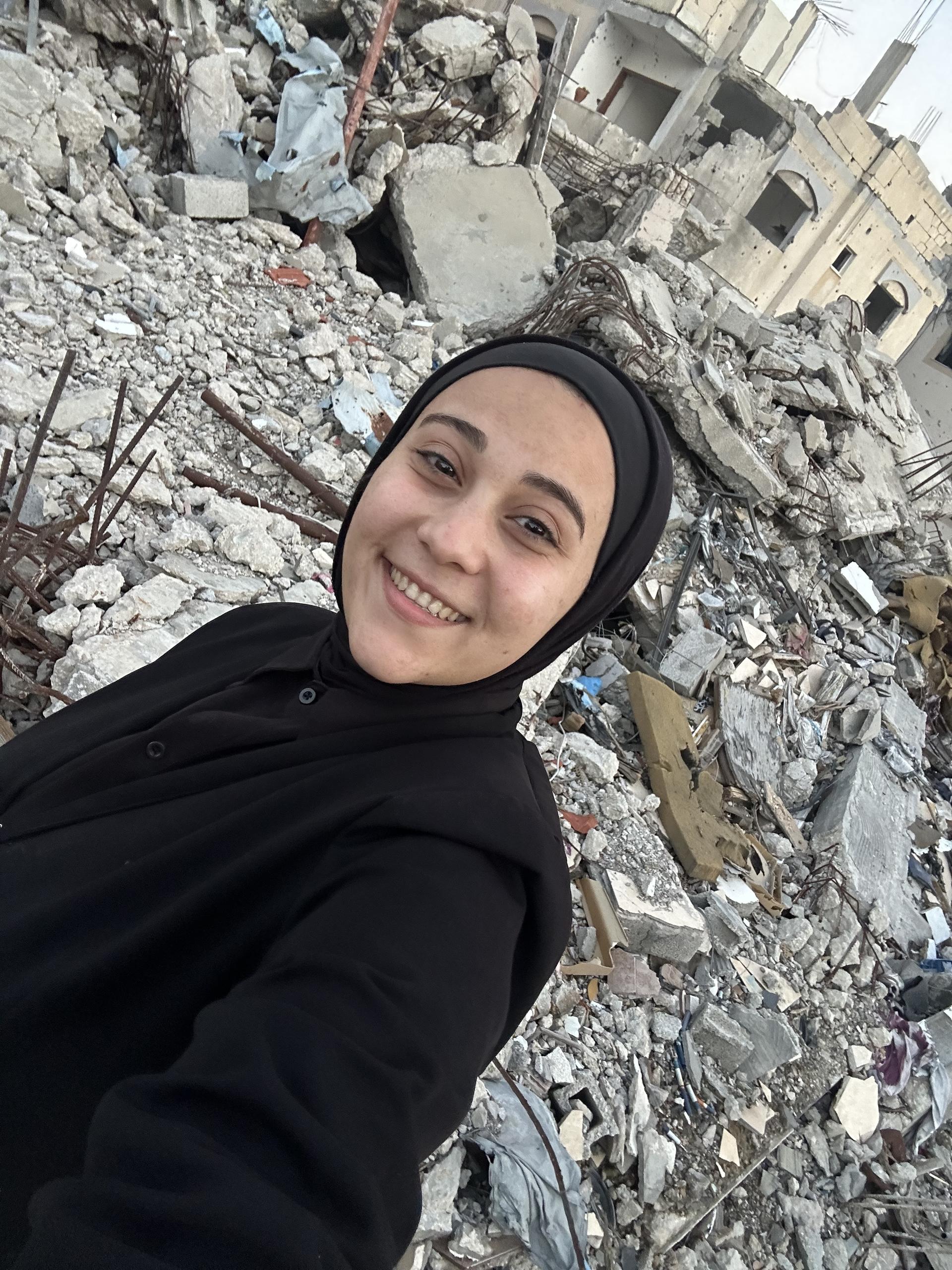 Photo: woman in a black hijab smiling amid rubble and ruined buildings.
